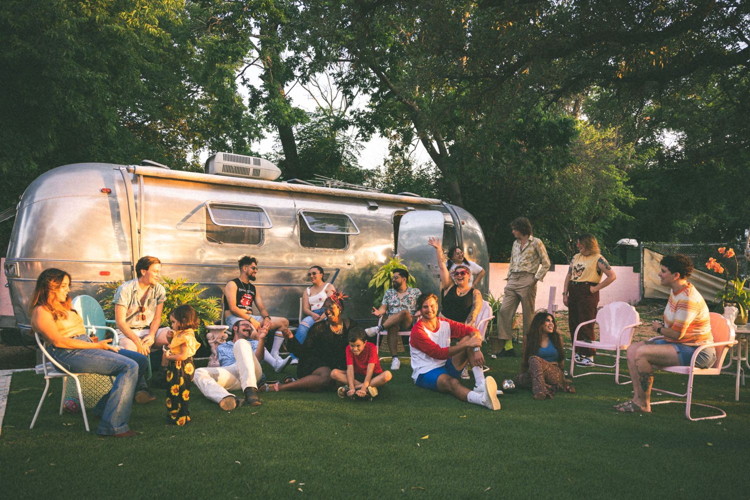 A group gathers outside around an Airstream trailer for a casual evening social gathering in a backyard setting.