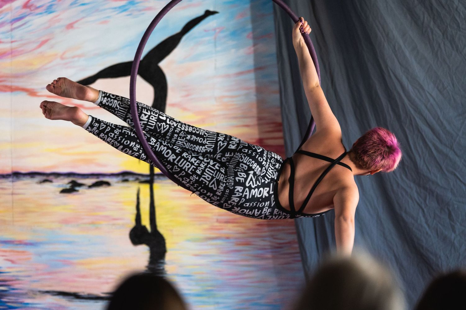 Aerial performer in patterned leggings performs on suspended equipment against colorful sunset backdrop.