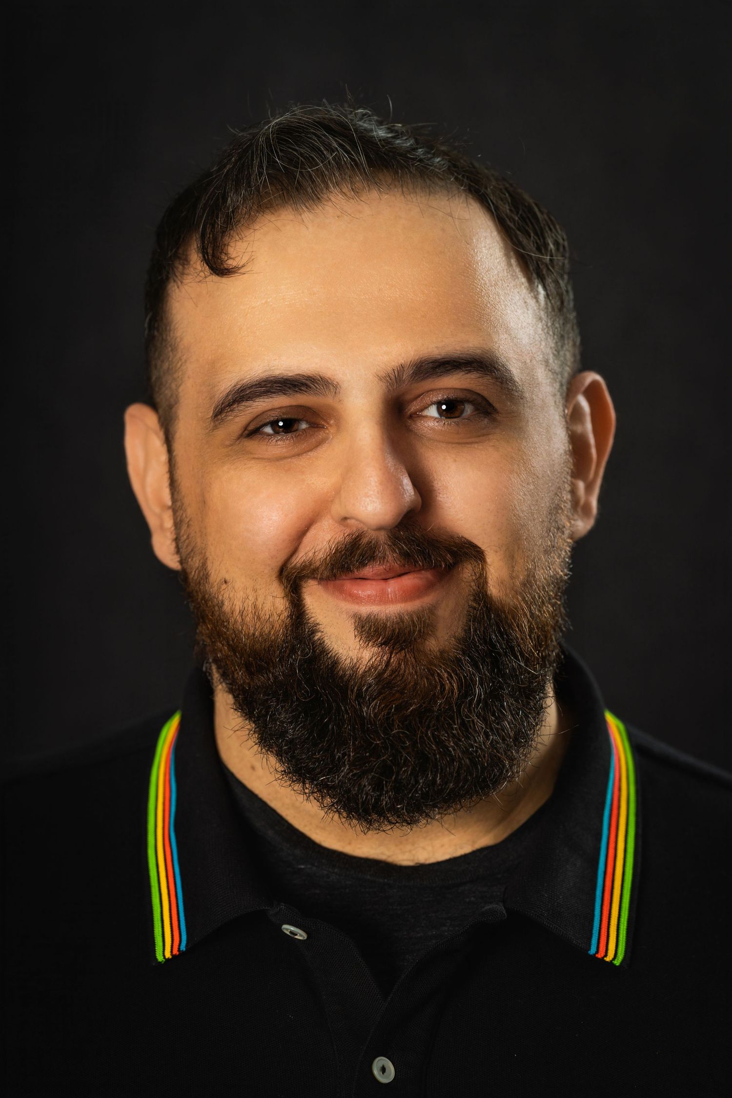 Professional studio portrait of person wearing black polo with rainbow stripes against dark background.