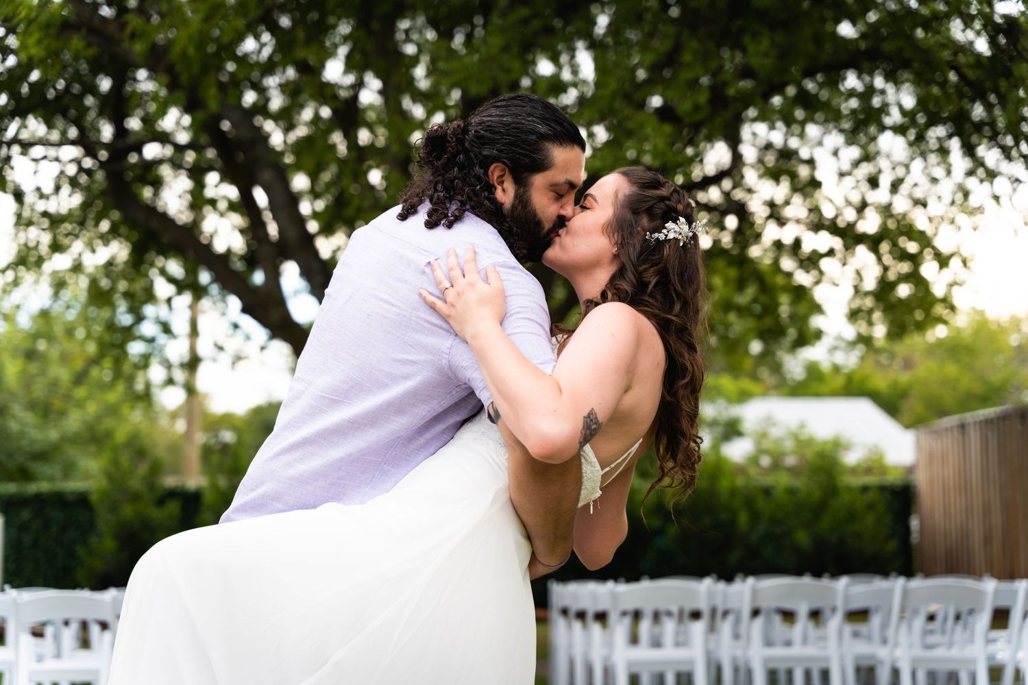 A romantic wedding kiss captured outdoors with white chairs set up for the ceremony.