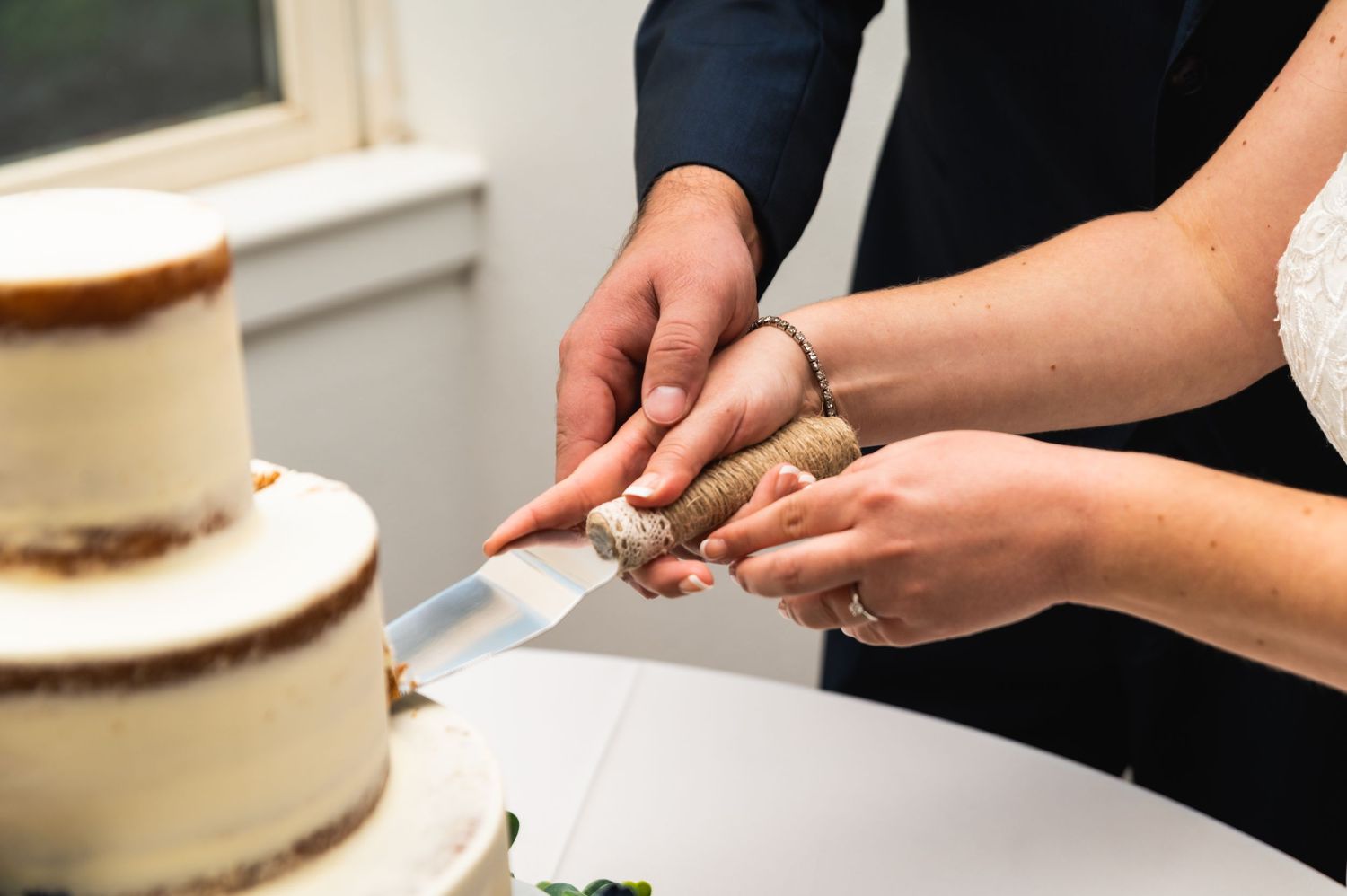 Close-up of hands cutting into a white wedding cake with a silver serving knife.