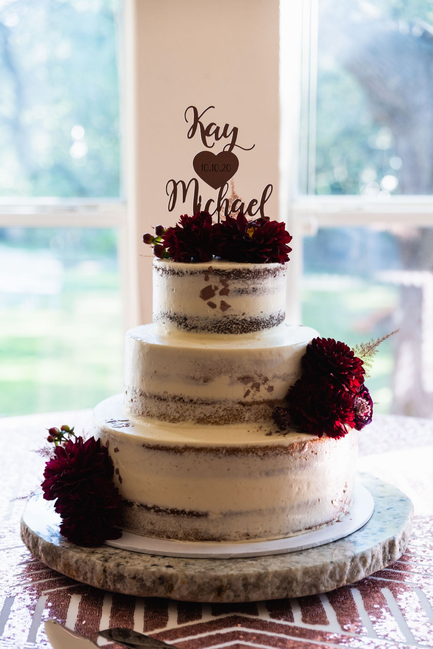 Three-tiered semi-naked wedding cake with burgundy flowers and custom name topper.