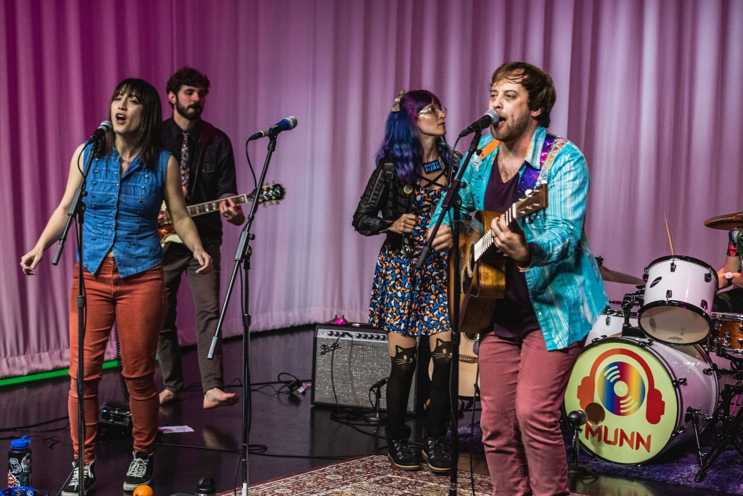 Folk band performs together on stage with colorful instruments and purple backdrop.