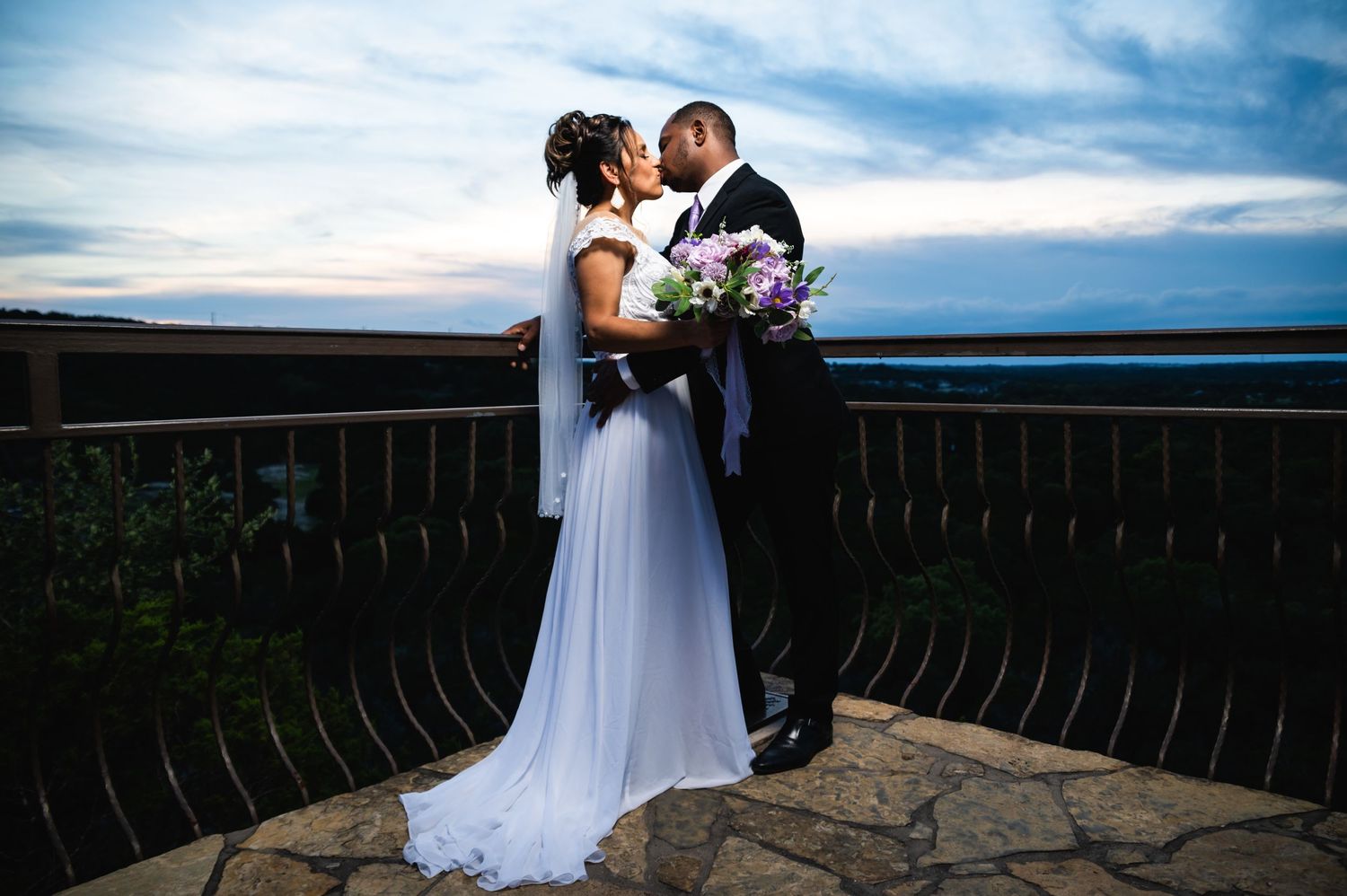 Romantic sunset portrait of newlyweds embracing on a stone balcony with mountain views.