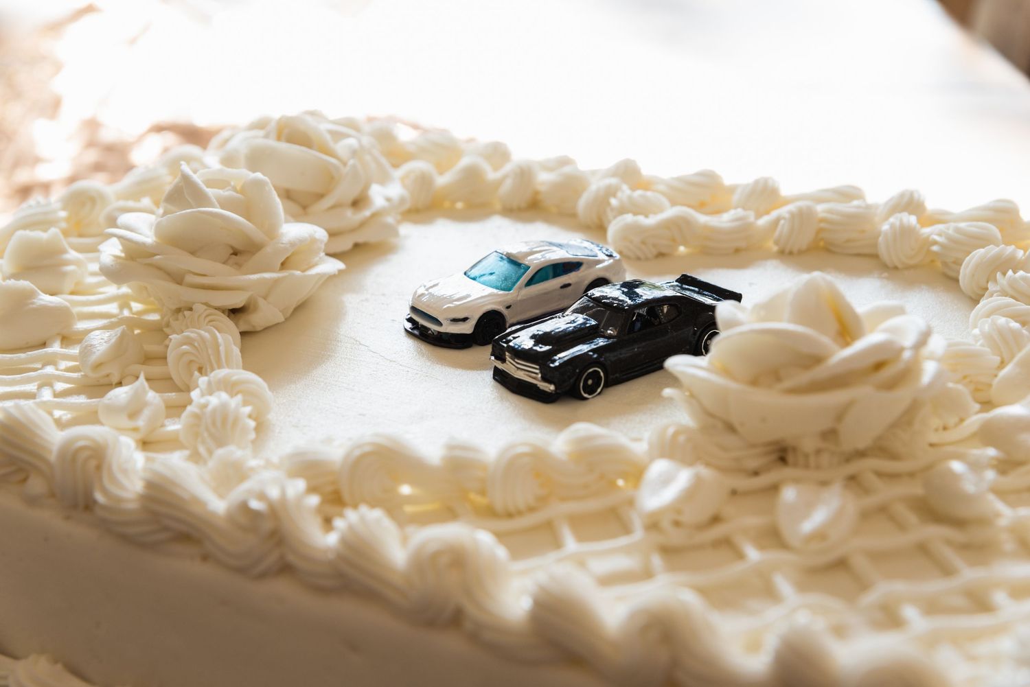 Two toy cars, one white and one black, sit atop a white frosted wedding cake.