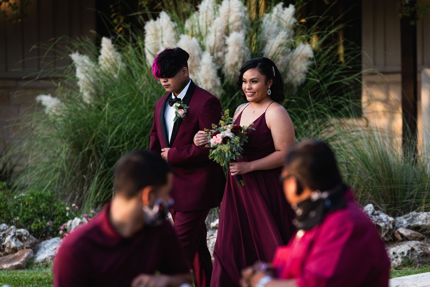 Wedding party members in burgundy attire gather near ornamental grasses.