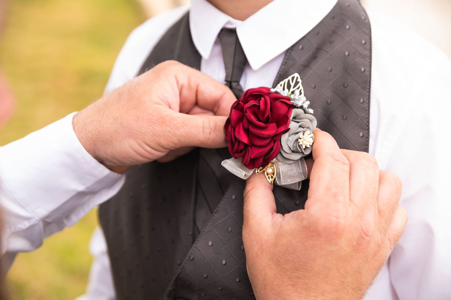 Close-up of hands pinning red rose boutonniere to grey vest.