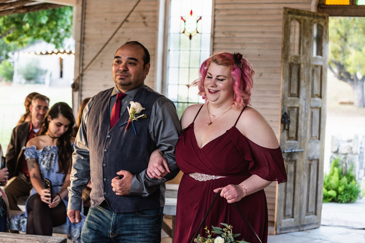 Wedding party members walking down the aisle in burgundy attire at a rustic chapel venue.