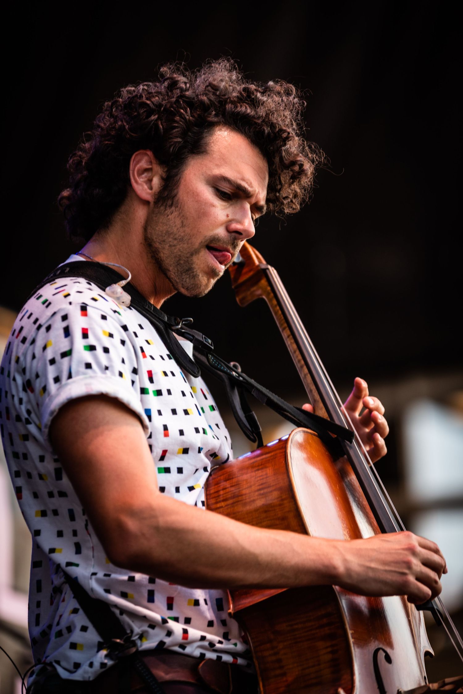 Cellist performing on stage wearing a white shirt with colorful geometric pattern.