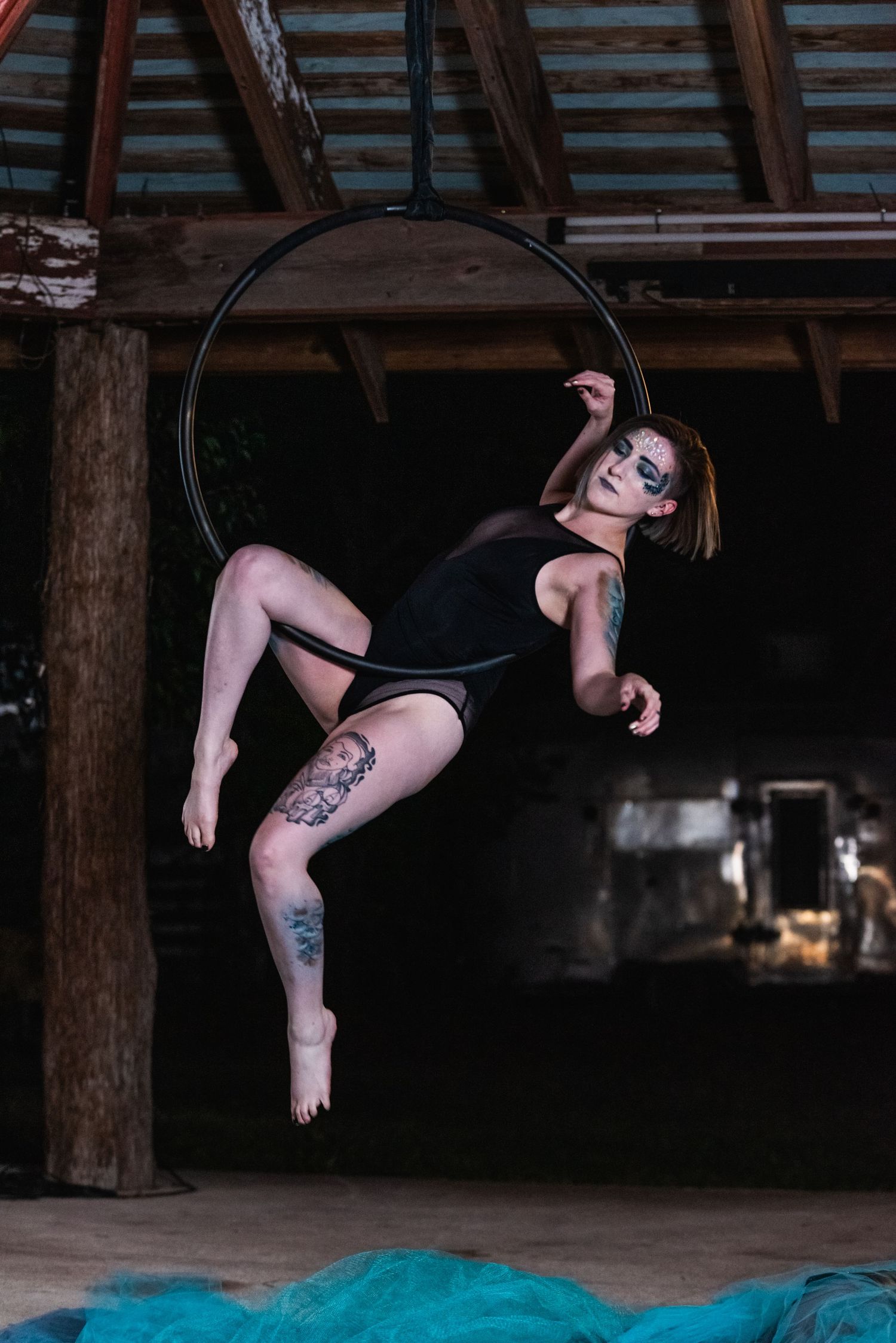 Tattooed aerial artist performs acrobatic pose on lyra hoop in rustic venue.