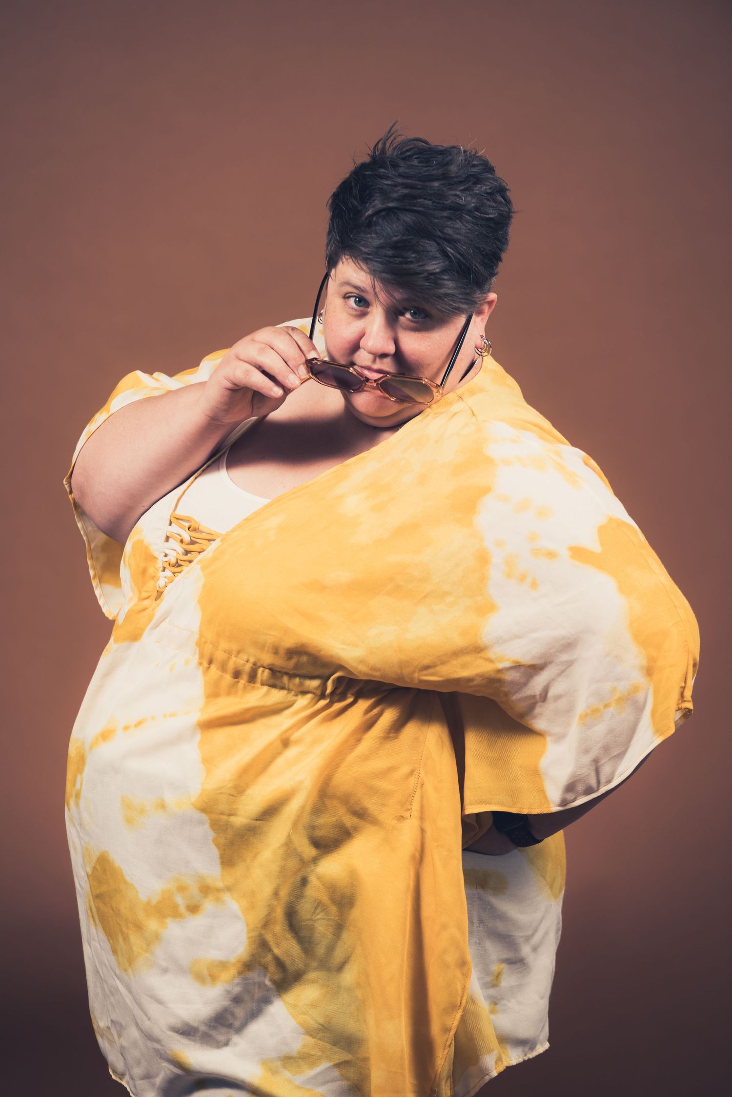 Playful portrait of person in yellow tie-dye outfit making silly expression against brown background.