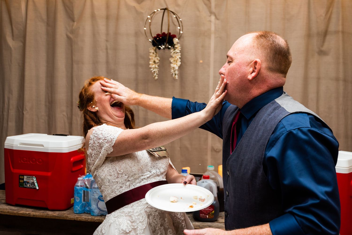 A playful cake feeding moment between a couple at their wedding reception with a red cooler in the background.