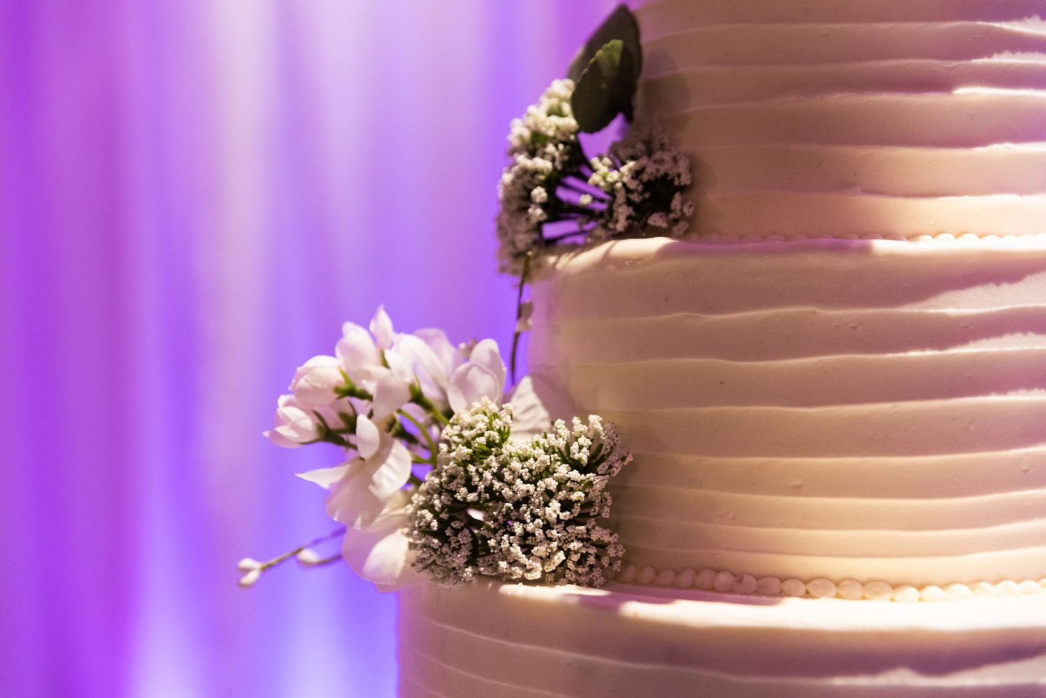 Close-up detail of white tiered wedding cake with floral decorations in purple lighting.