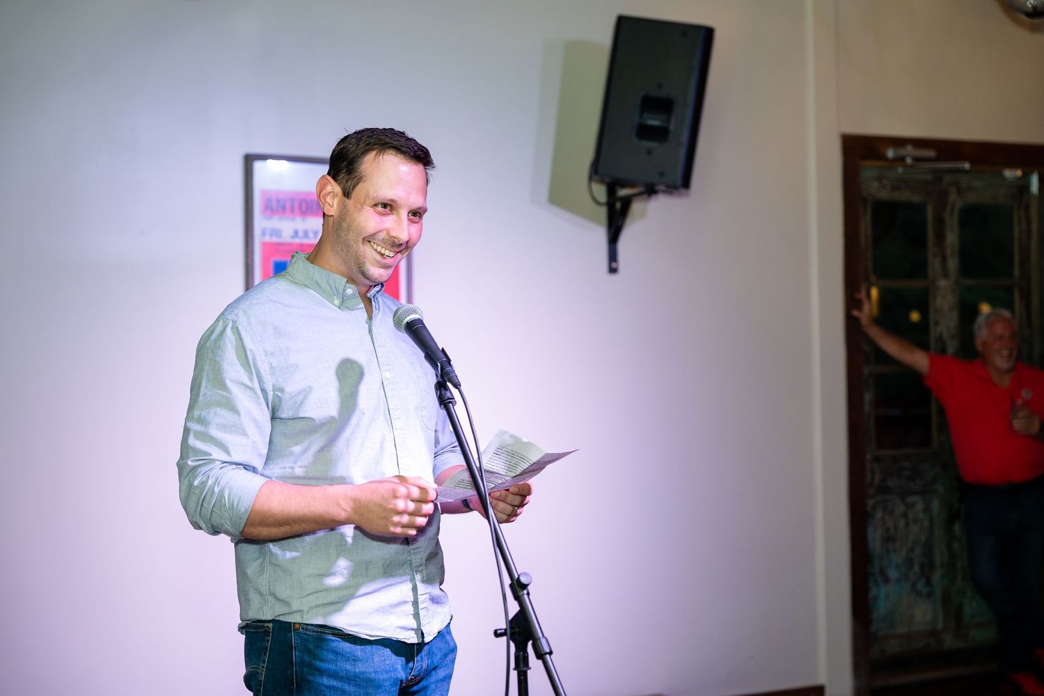 A speaker addresses an audience from a microphone stand in a dimly lit venue.