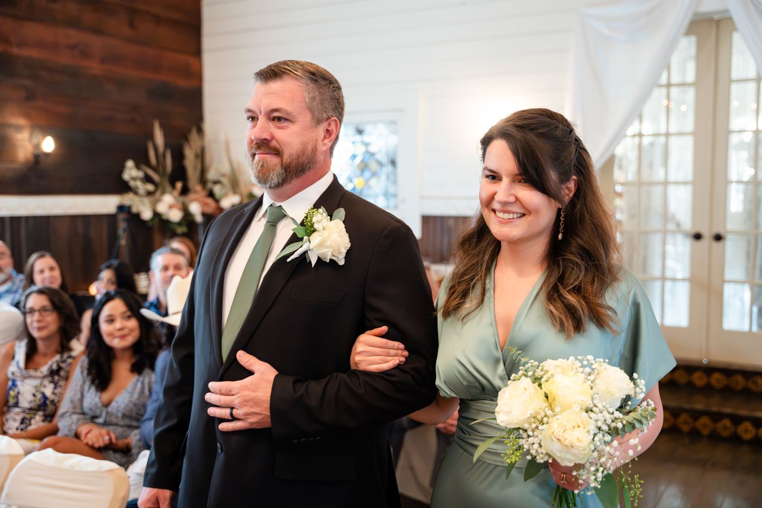 Wedding party members walking down the aisle in sage green attire with white floral bouquets.