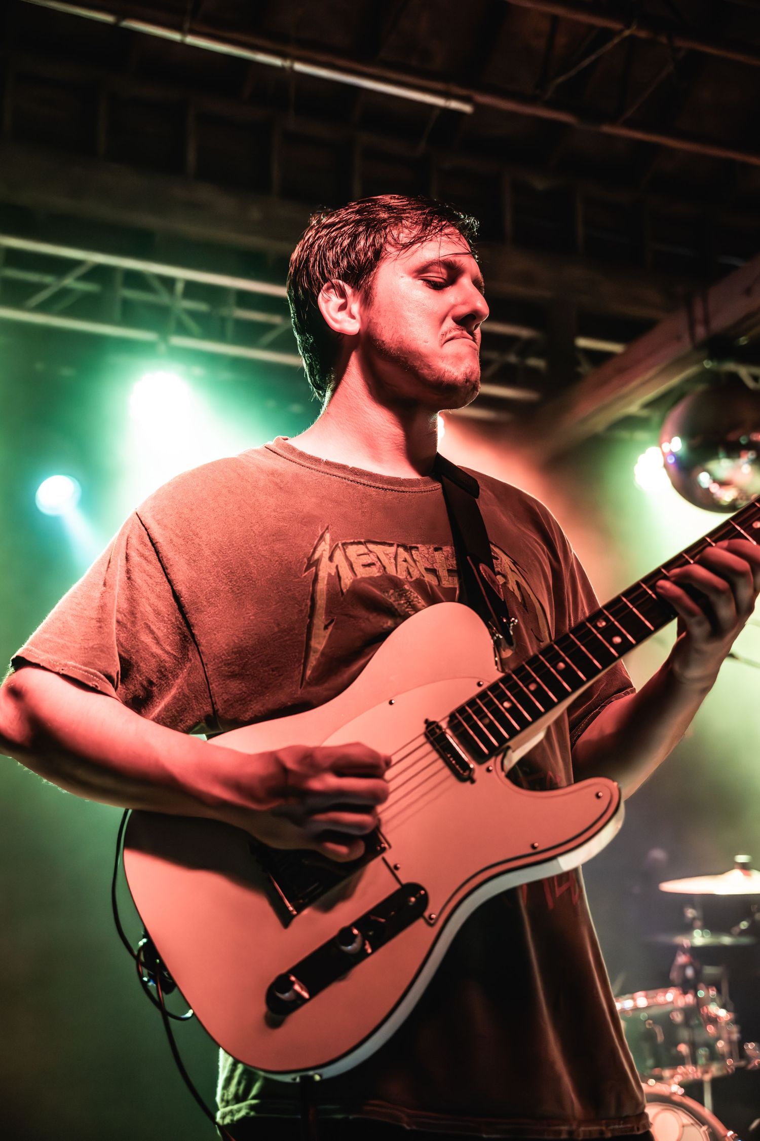 Guitarist plays light colored electric guitar on dimly lit stage with green backlighting.