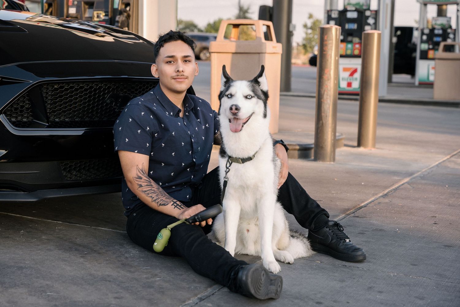 Person with husky dog sitting by black Mustang at gas station.