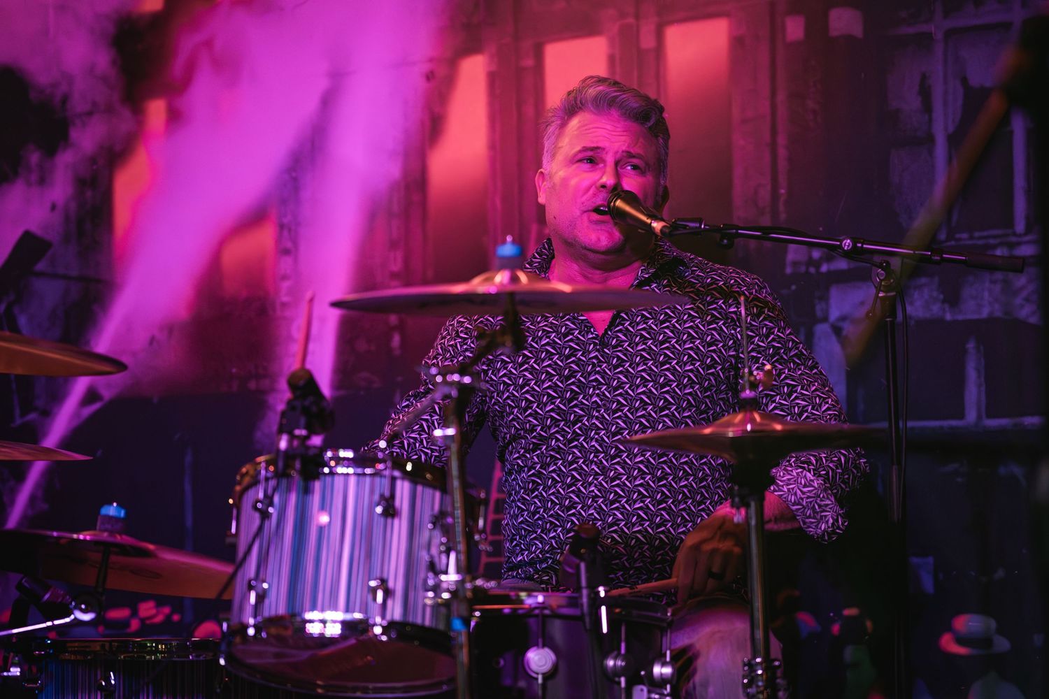 Musician plays drums in a patterned shirt under dramatic pink stage lighting.
