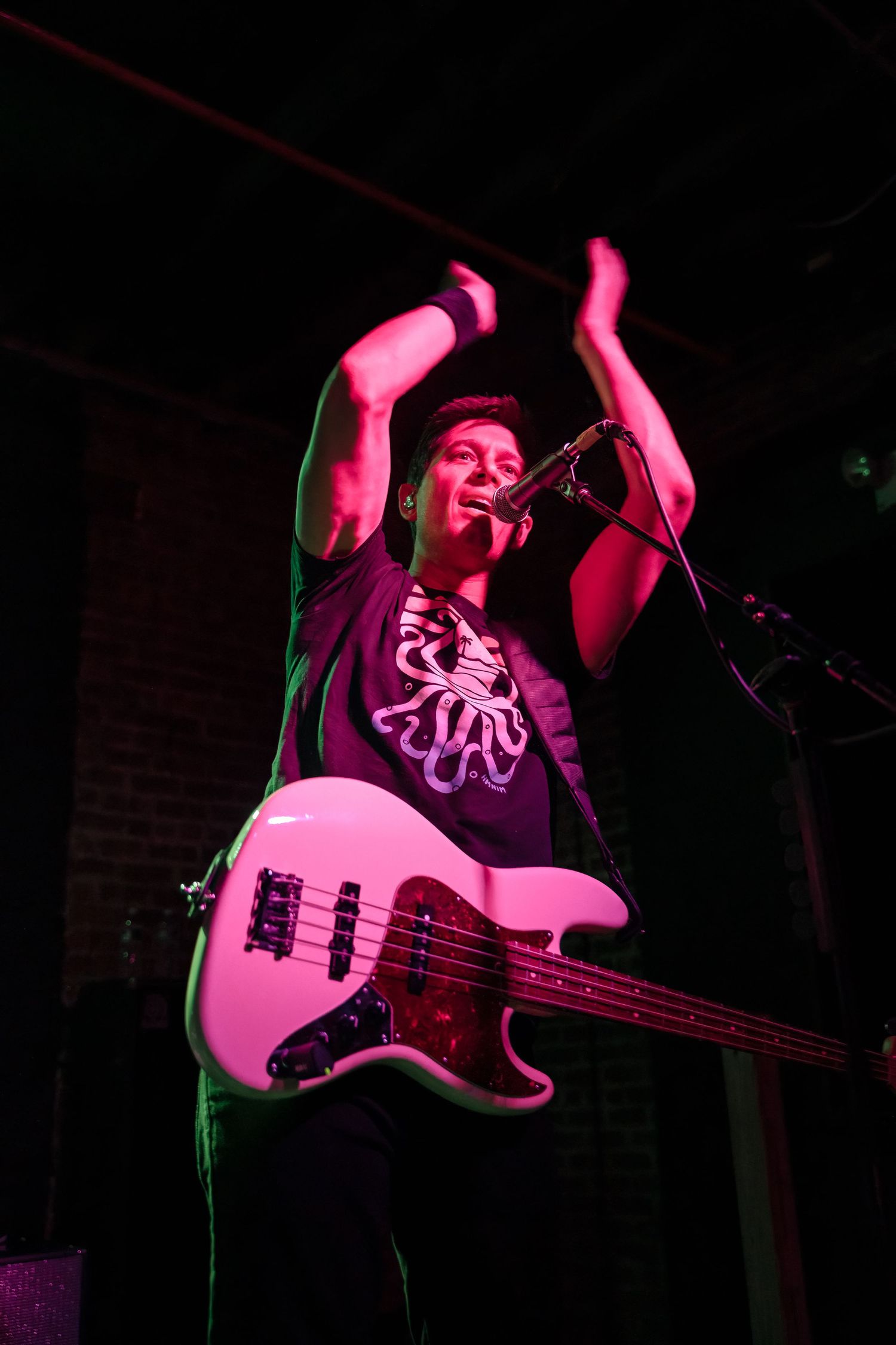 Musician plays pink bass guitar while singing passionately under dramatic stage lighting.