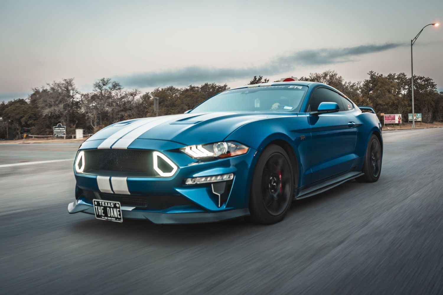 A blue Ford Mustang with white racing stripes speeding on a road during golden hour.