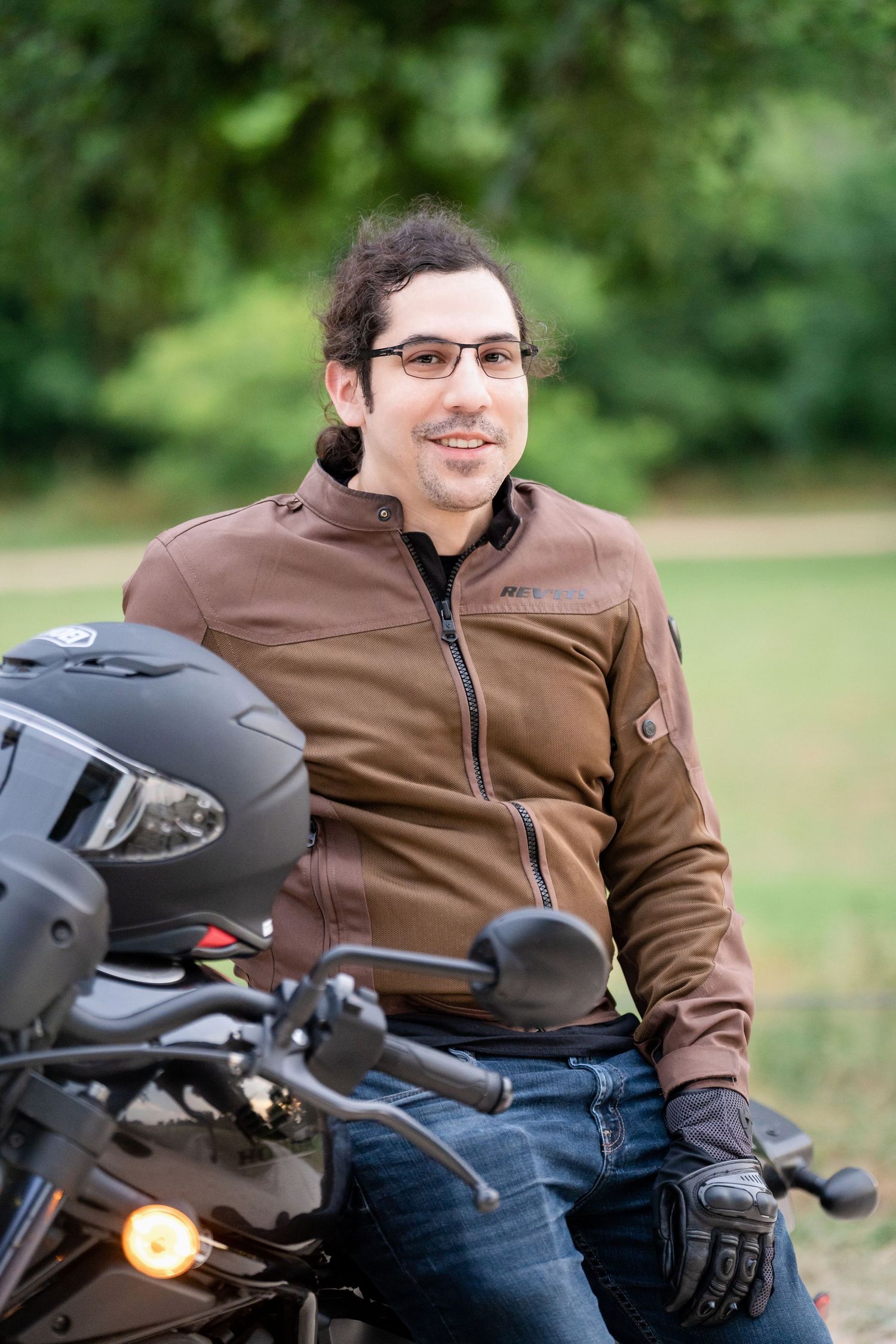A motorcyclist in brown jacket and gloves sits on their parked motorcycle in a park setting.
