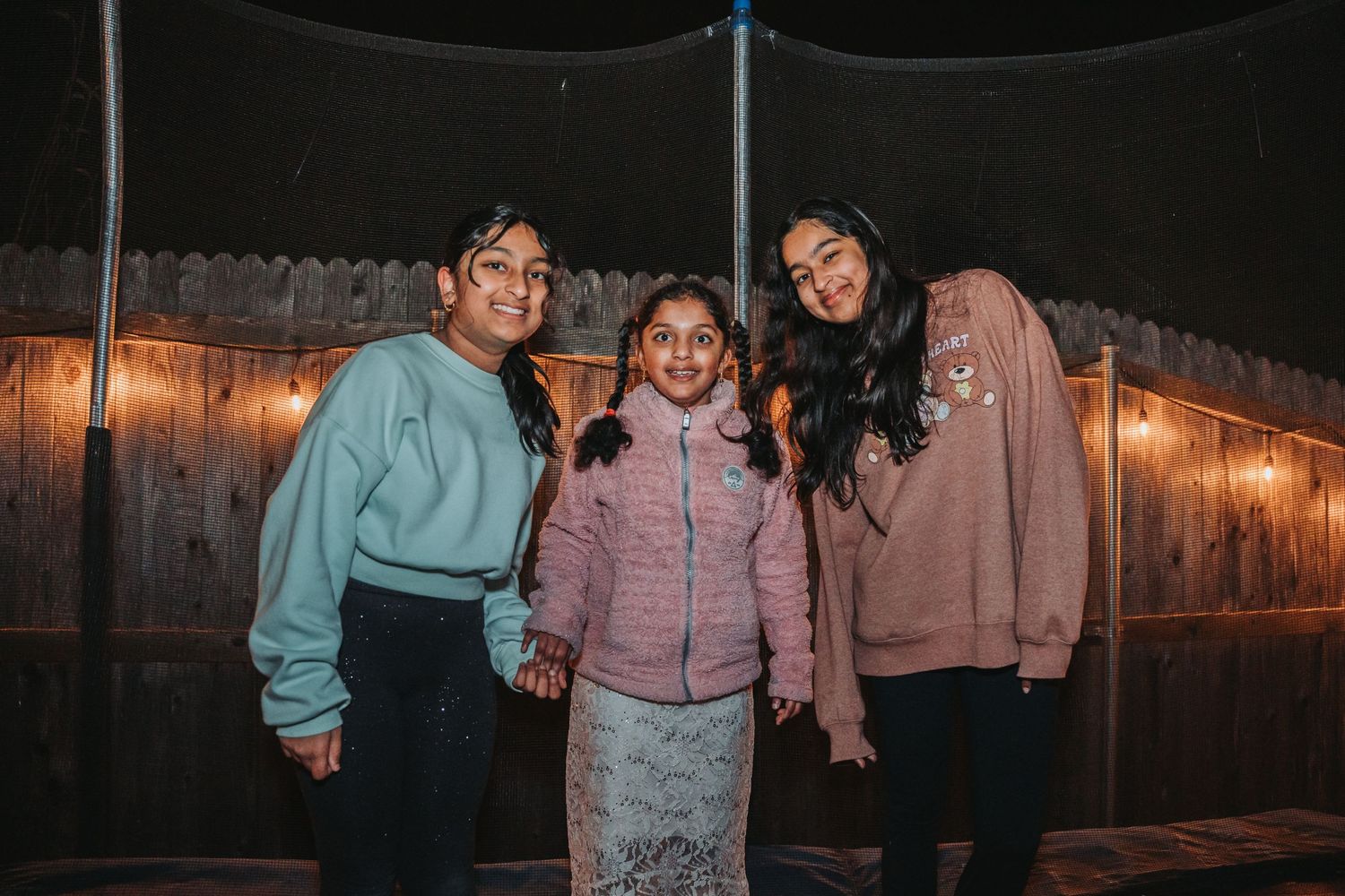 Friends posing together outdoors at night in front of a wooden fence with string lights.