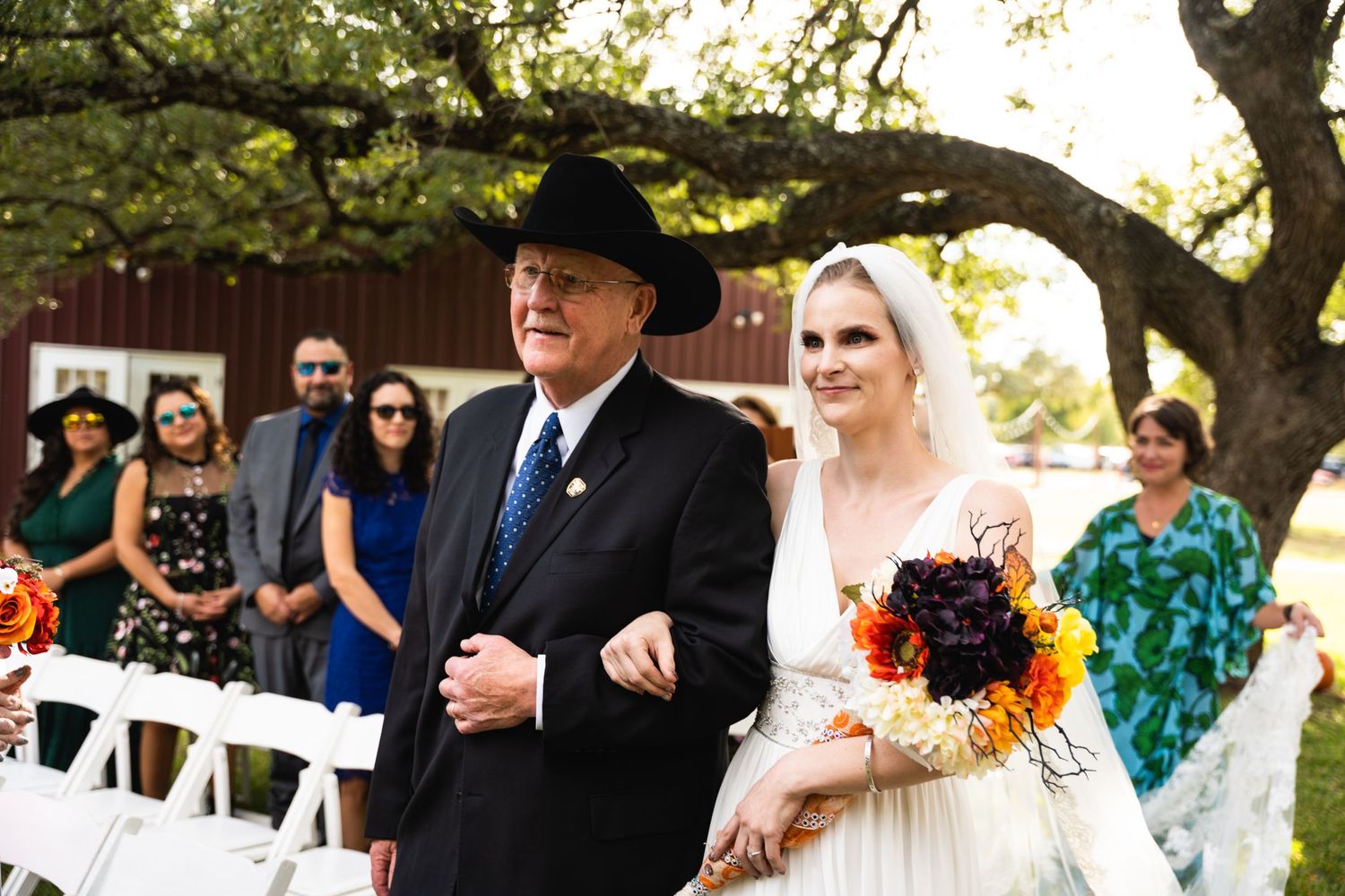 A wedding processional moment under a tree with guests watching from white chairs.