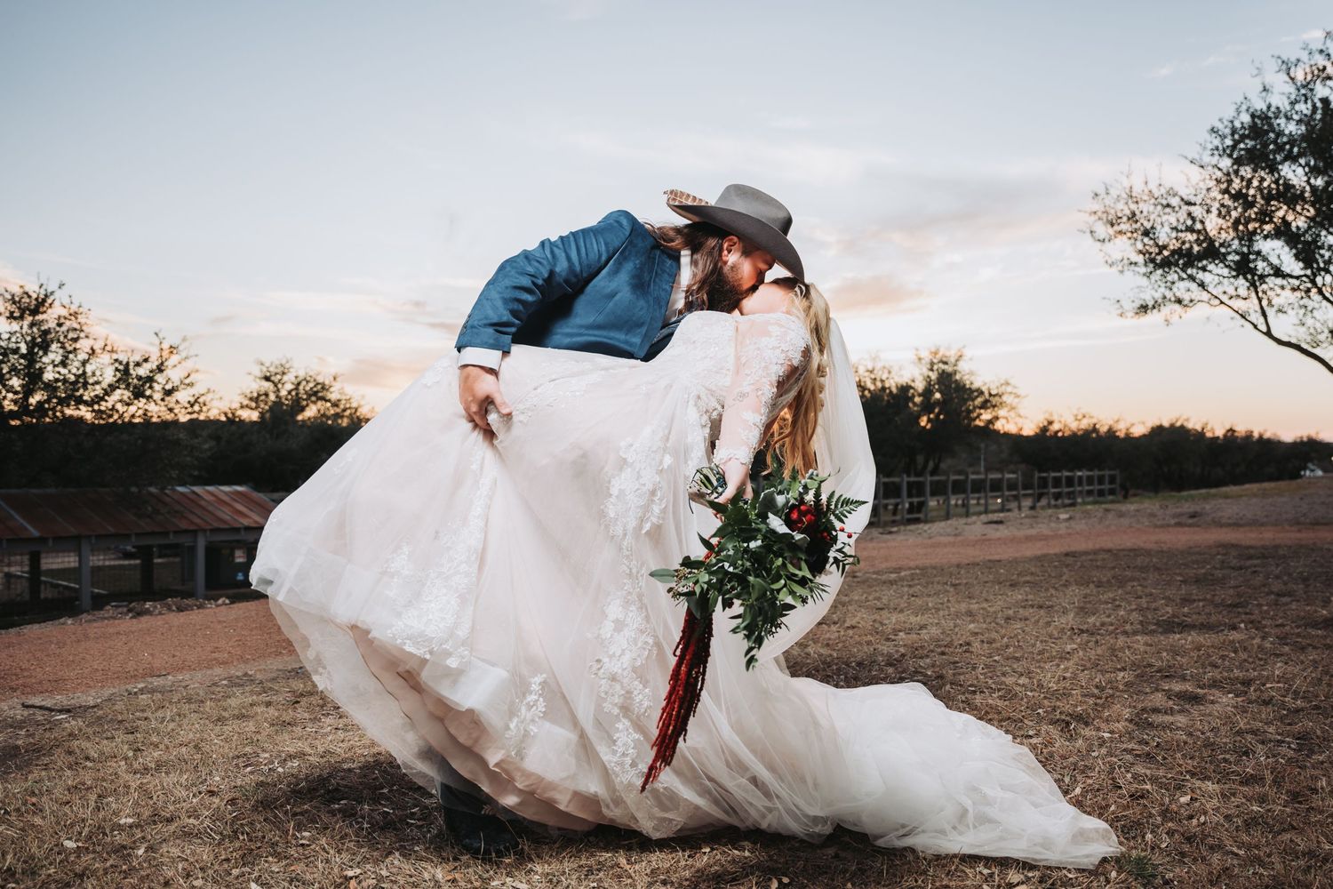 A romantic dip kiss at sunset with the bride's dress flowing dramatically in a rustic outdoor setting.