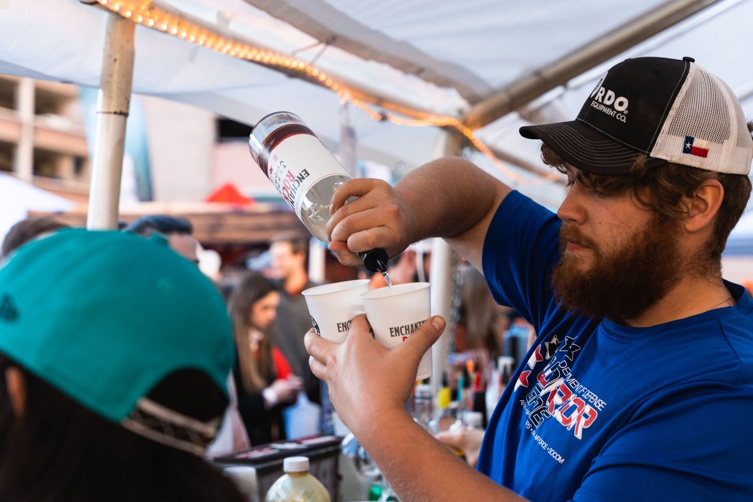 Person pouring drinks at an outdoor event wearing a baseball cap and blue shirt.