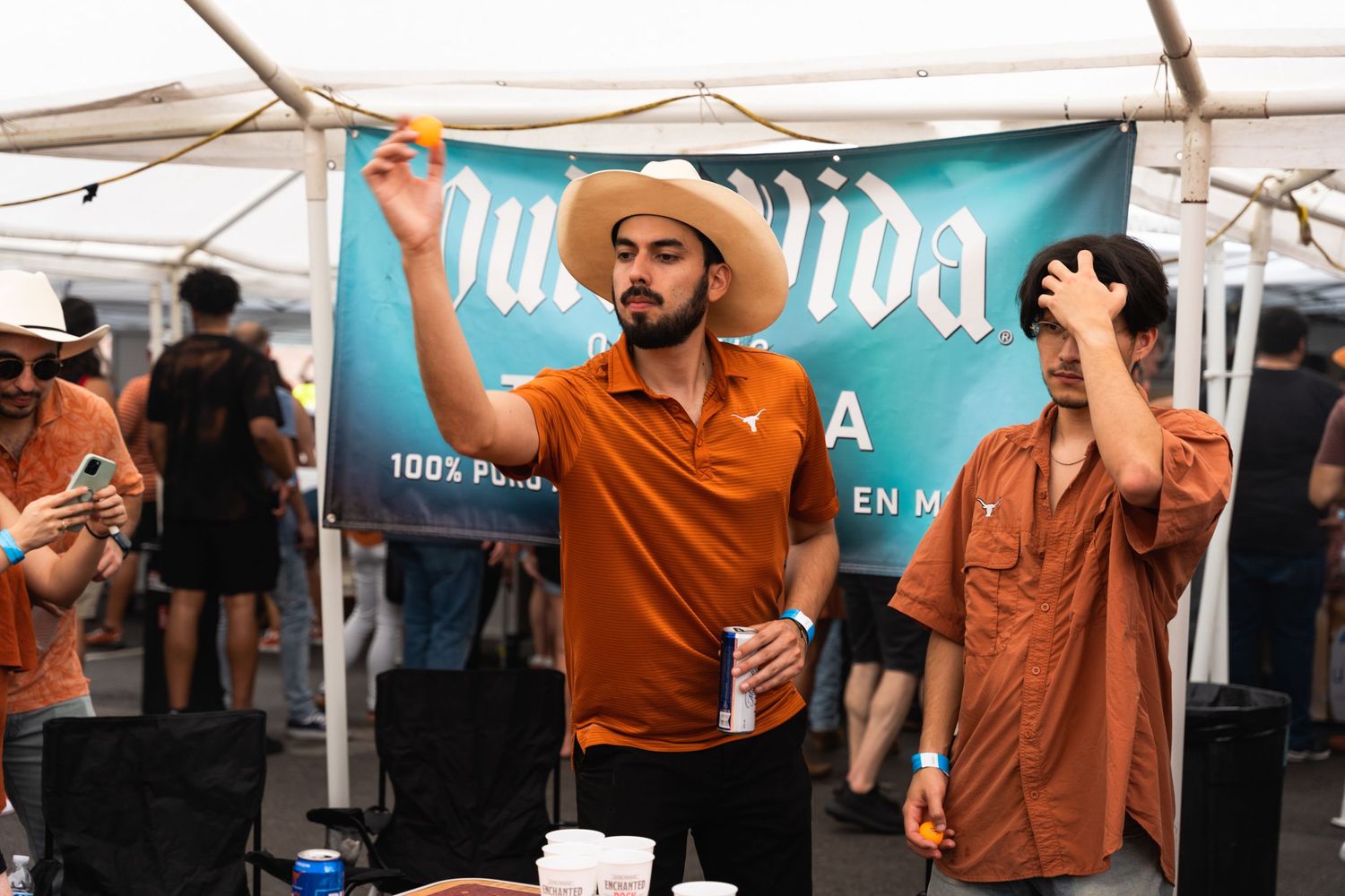 Two people in orange and brown shirts celebrating under a tent banner at an outdoor event.