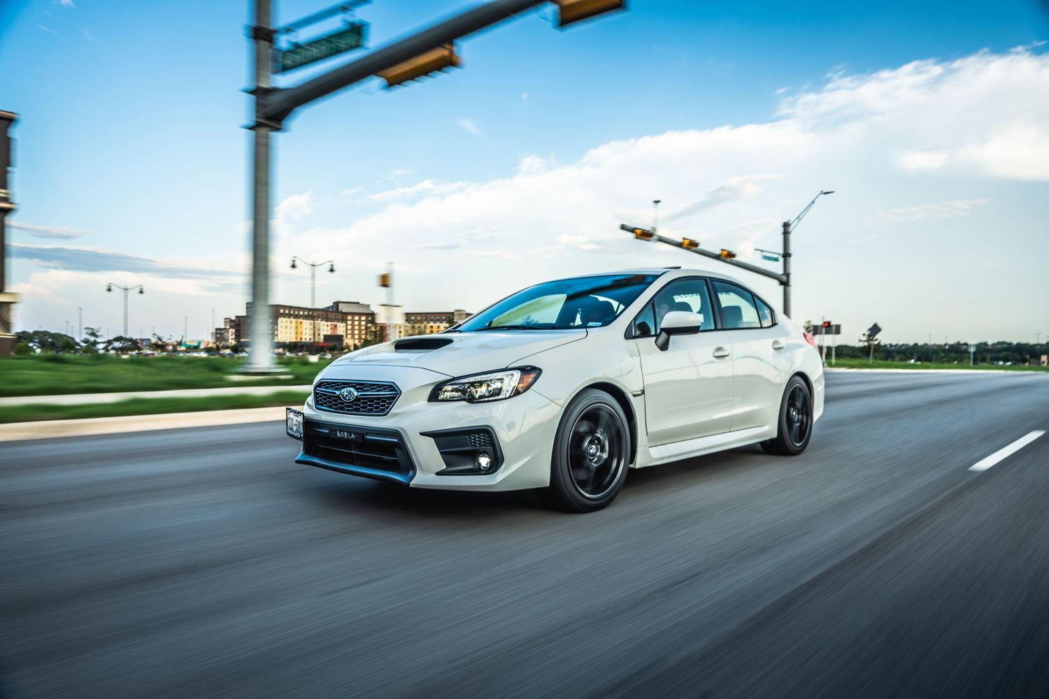 Modified white Subaru WRX sedan speeding on a city street with traffic lights visible.