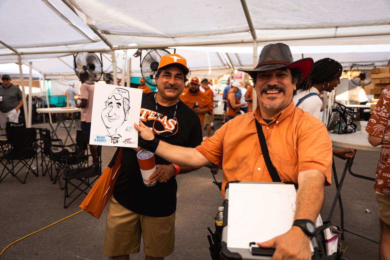 Two people pose with caricature artwork at an outdoor event under a white canopy.