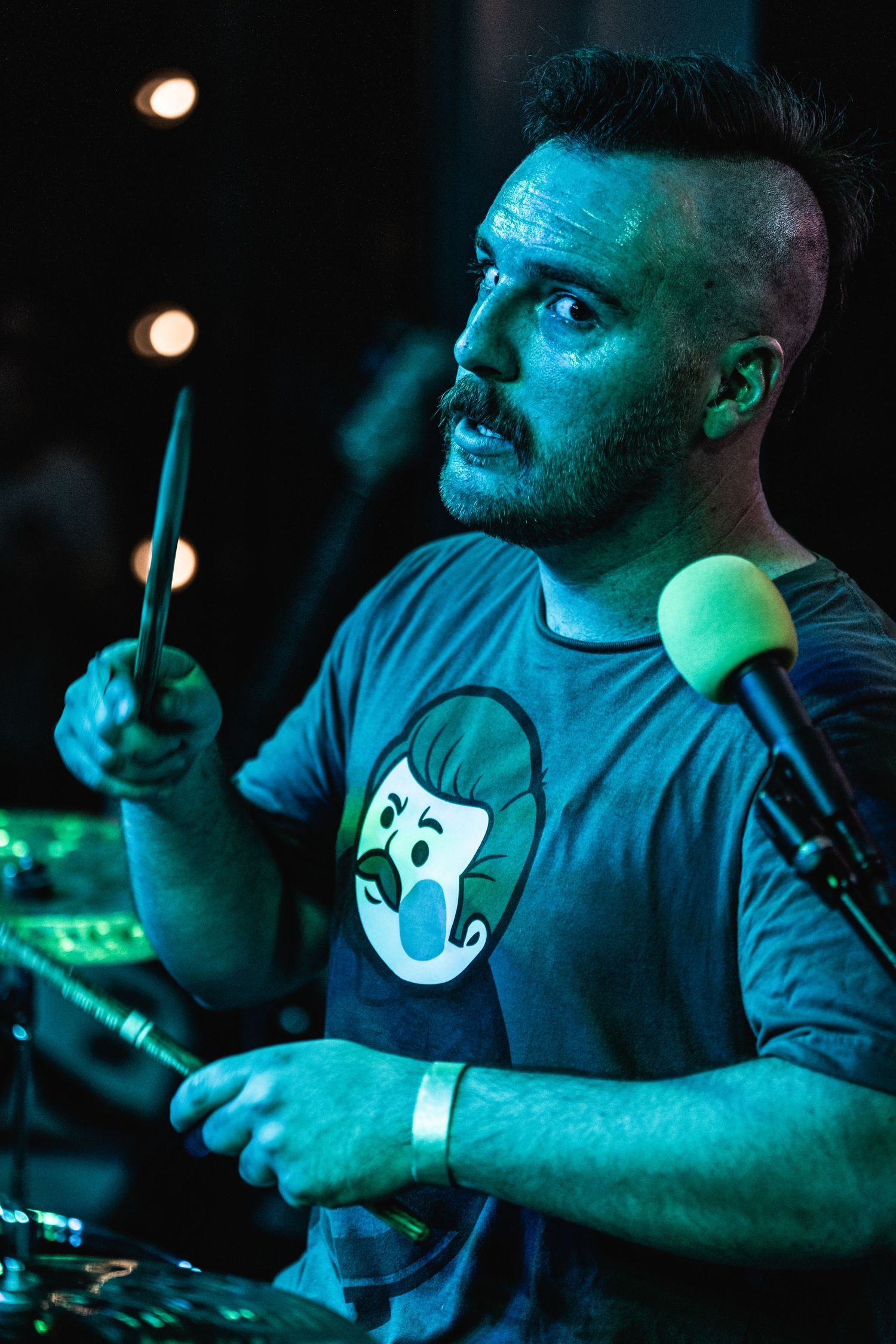Drummer performing with drumsticks under cyan stage lighting while wearing graphic t-shirt.