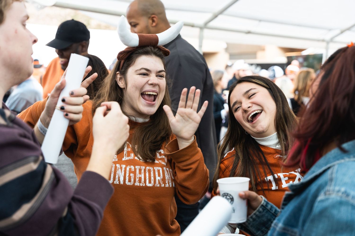 Energetic crowd of students celebrating and cheering at a university event under a white tent.