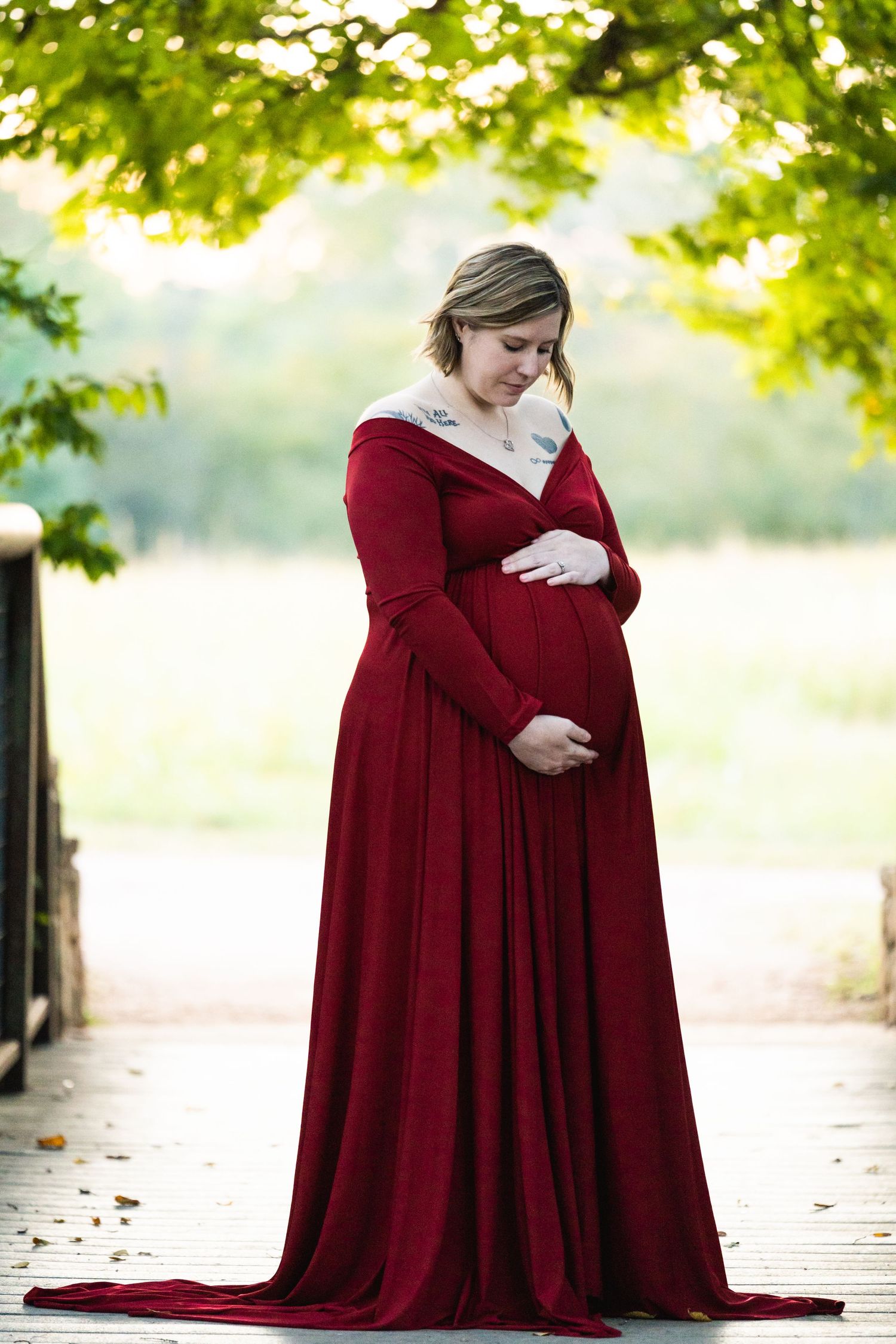 A pregnant woman in a flowing burgundy dress poses outdoors under green foliage on a garden path.