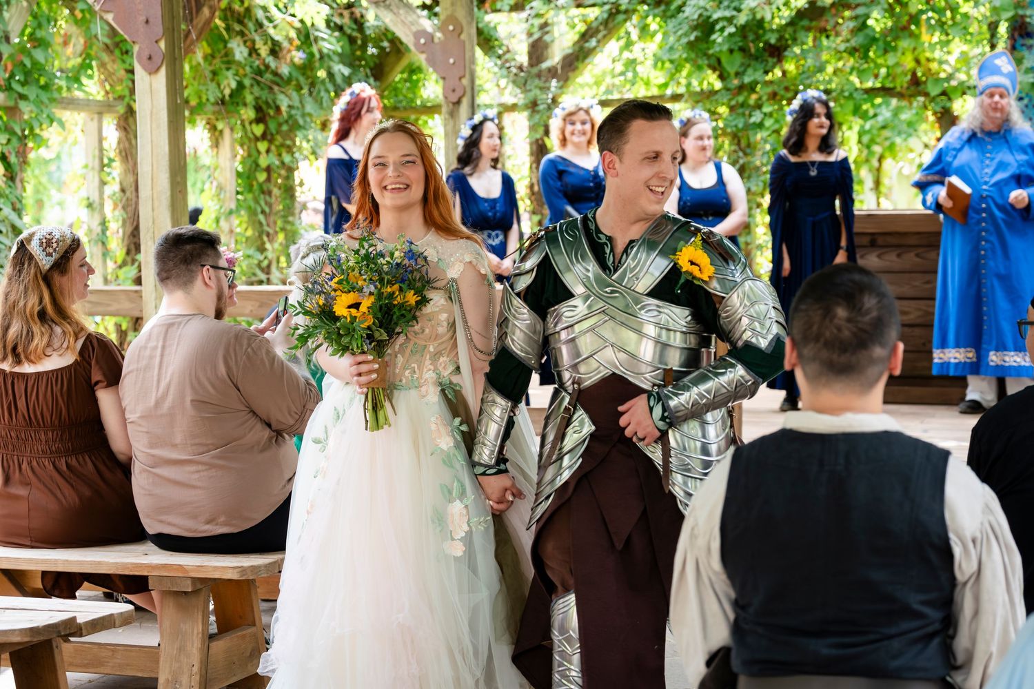Wedding celebration scene with guests gathered outdoors under green foliage and decorative lighting.