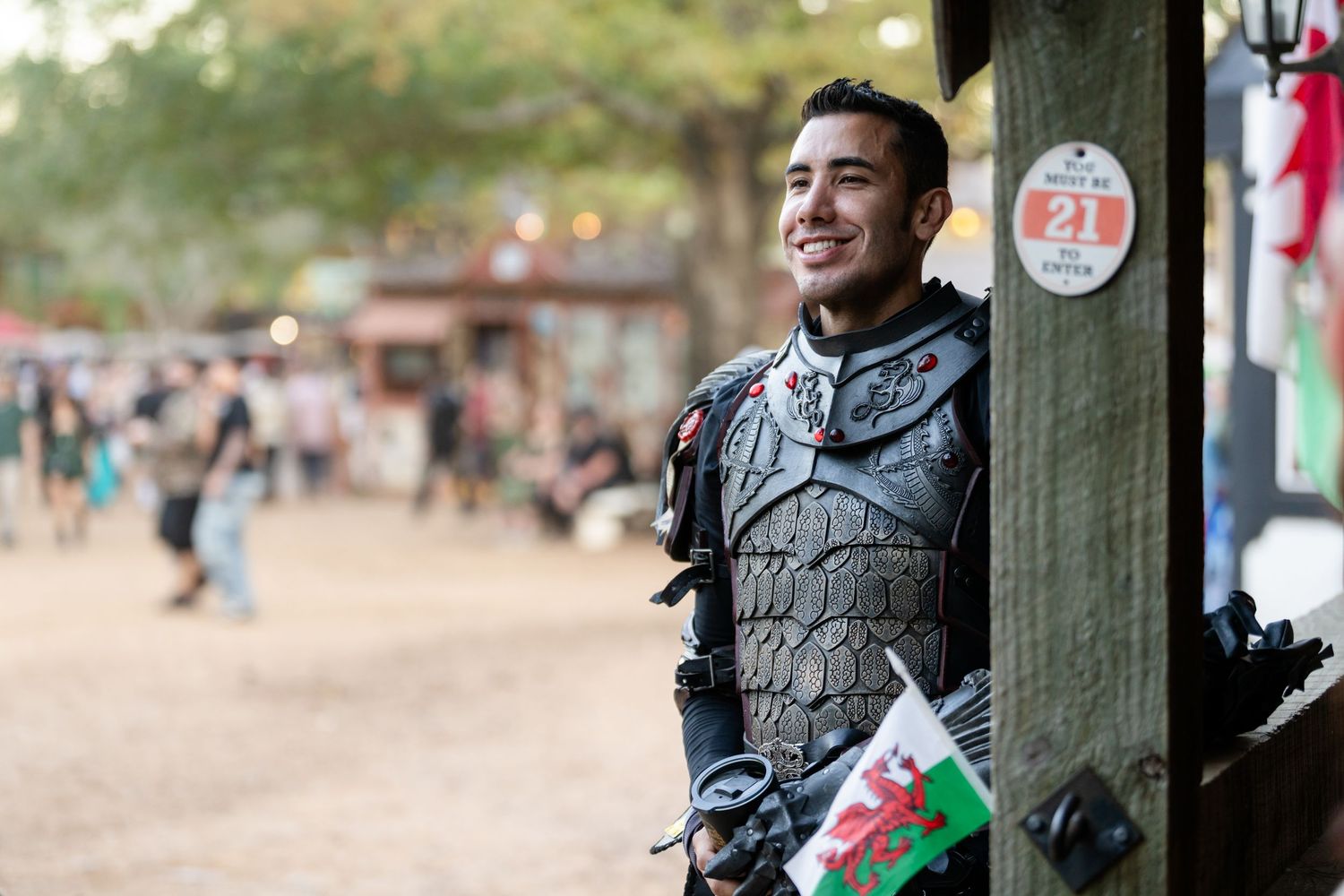 A performer in medieval armor stands smiling at a renaissance festival while holding a Welsh flag.