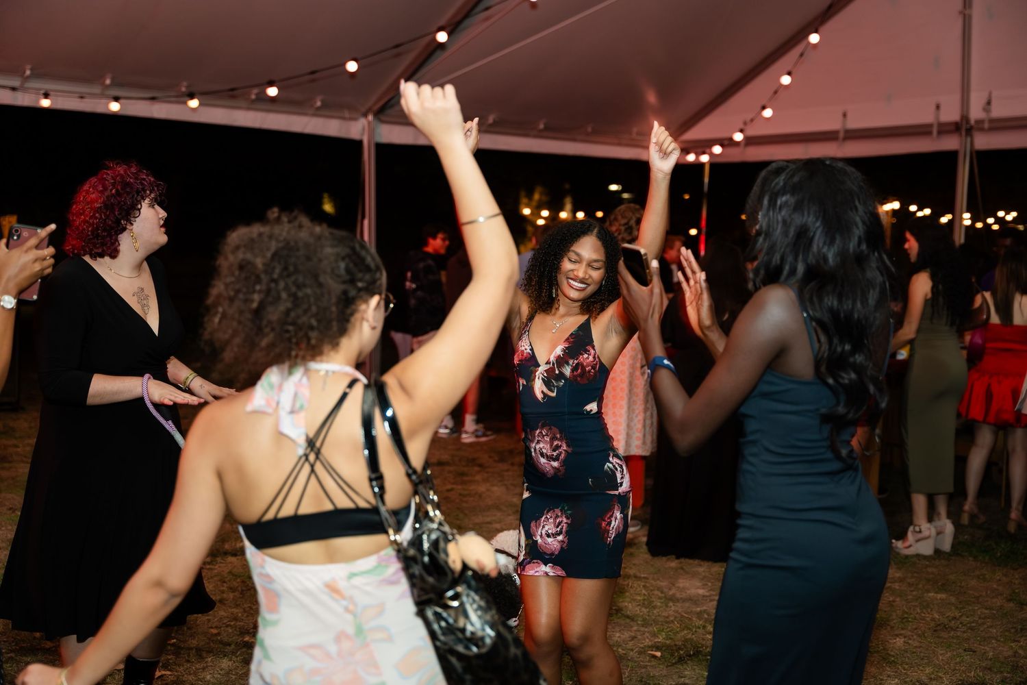 People dancing and celebrating at a nighttime event under a tent with string lights overhead.
