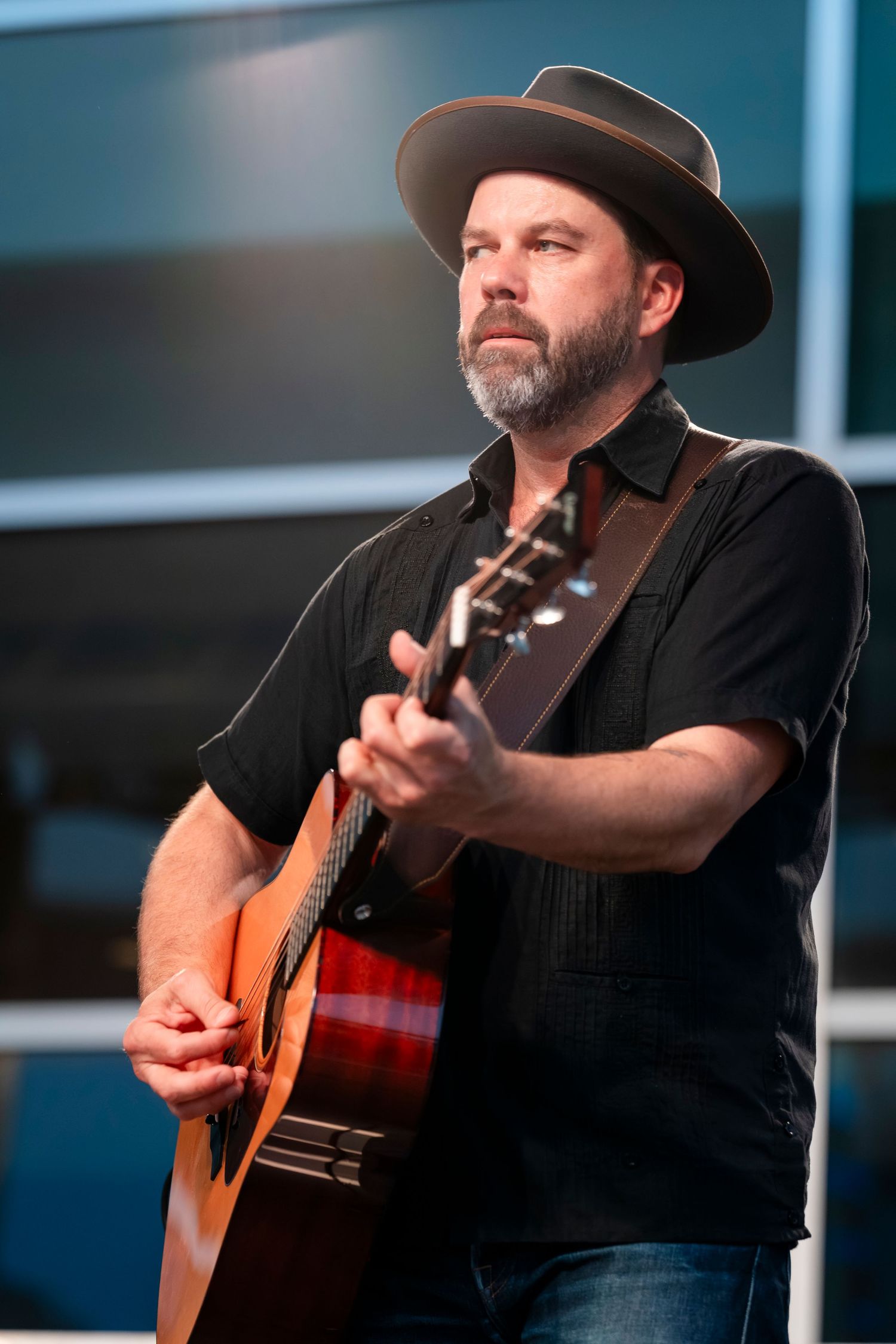 A musician in a black shirt and brown hat plays acoustic guitar on stage during a performance.