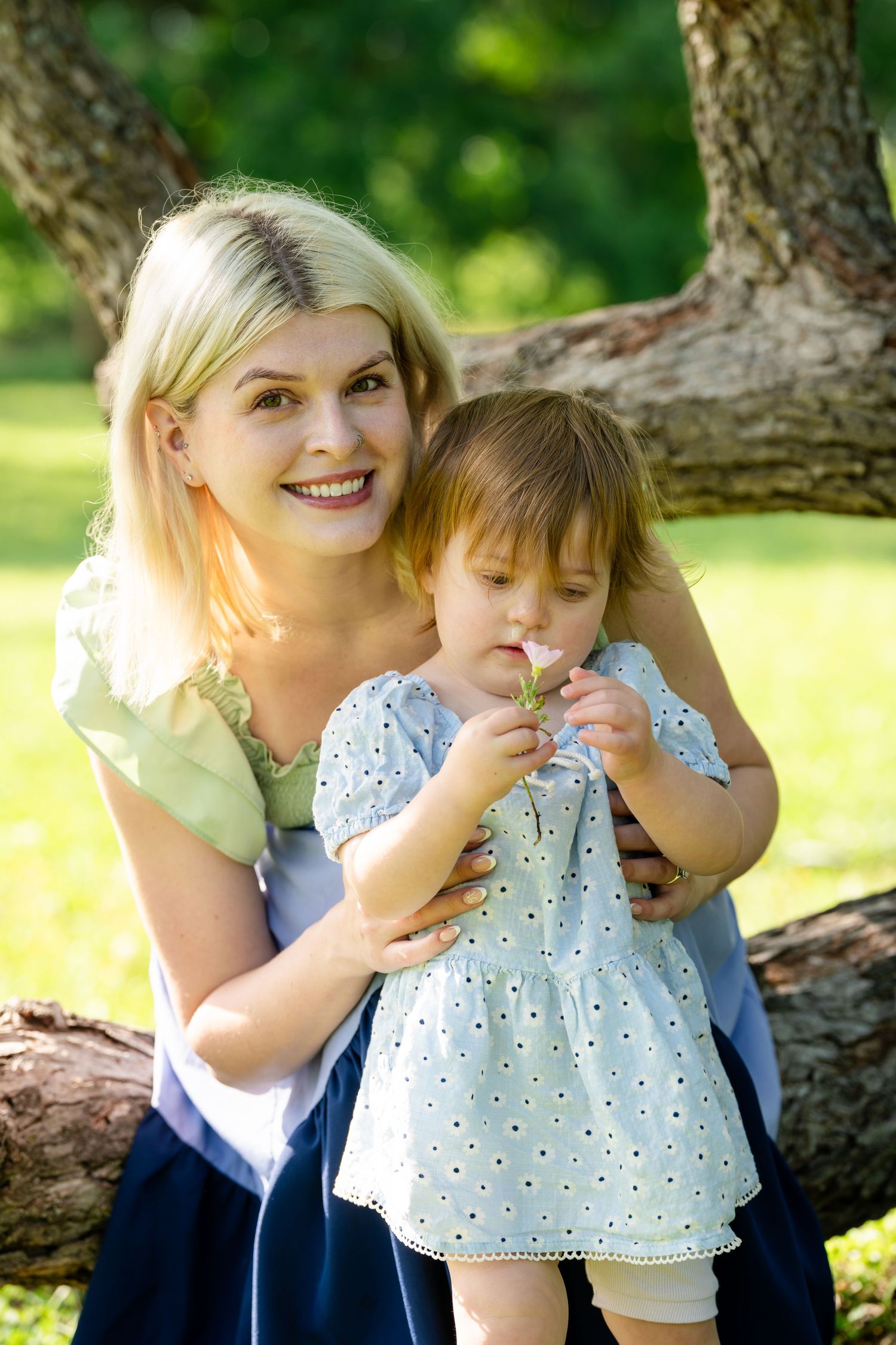 Smiling blonde mother hugging toddler girl in polka dot dress smelling a flower in a sunny green park.