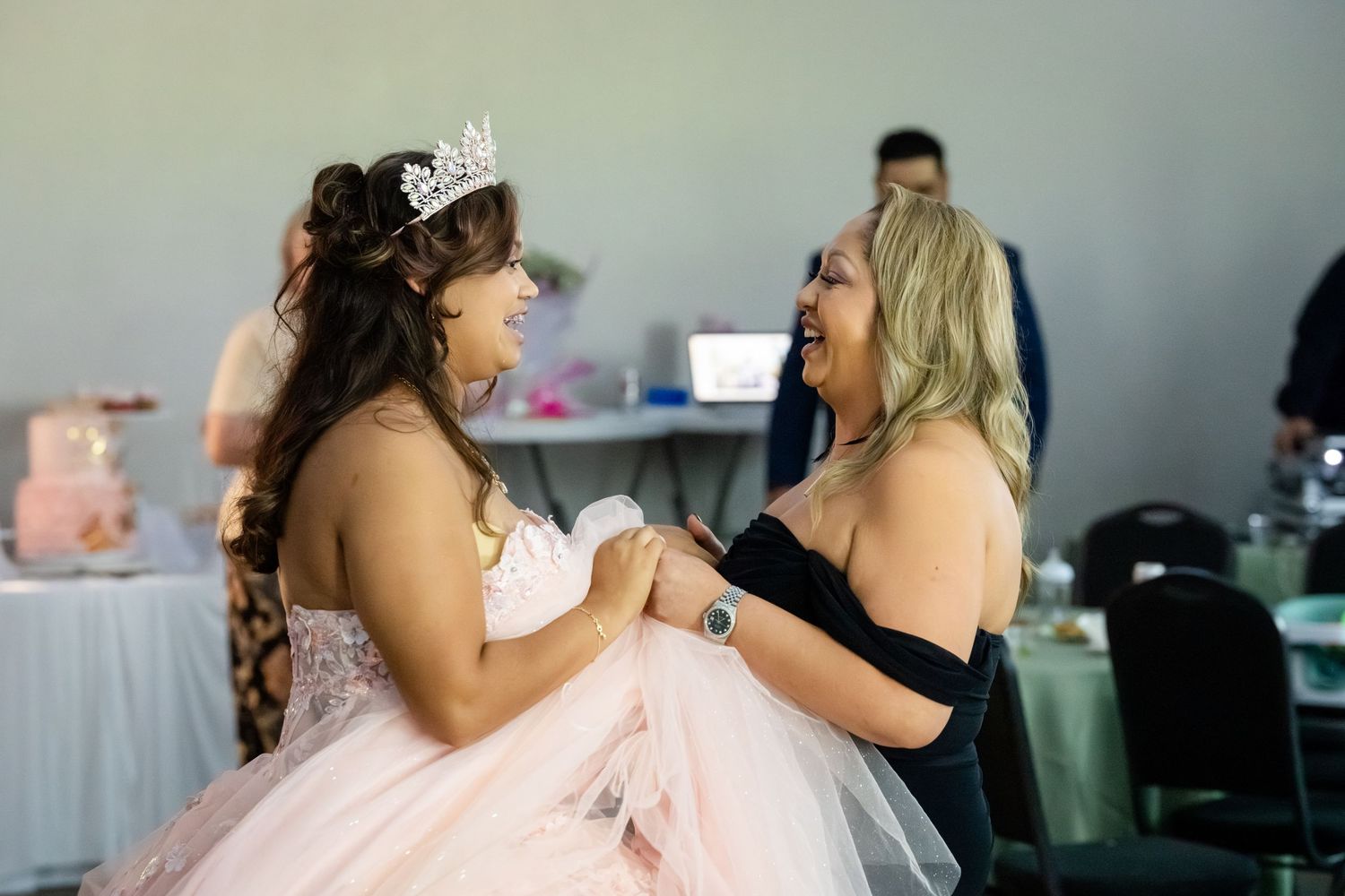 A young woman in a pink quinceañera gown and tiara joyfully dances with an older woman in a black dress at a celebration.