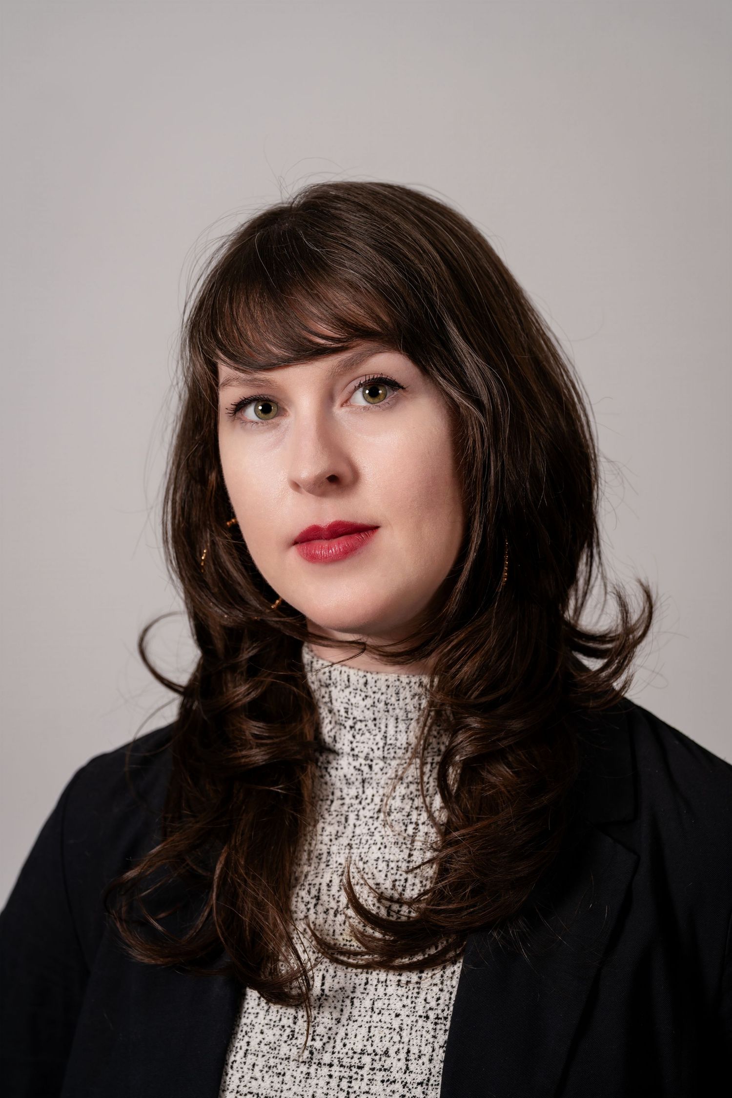 Professional headshot of a young woman with dark brown hair, red lips, wearing a black blazer and white turtleneck.