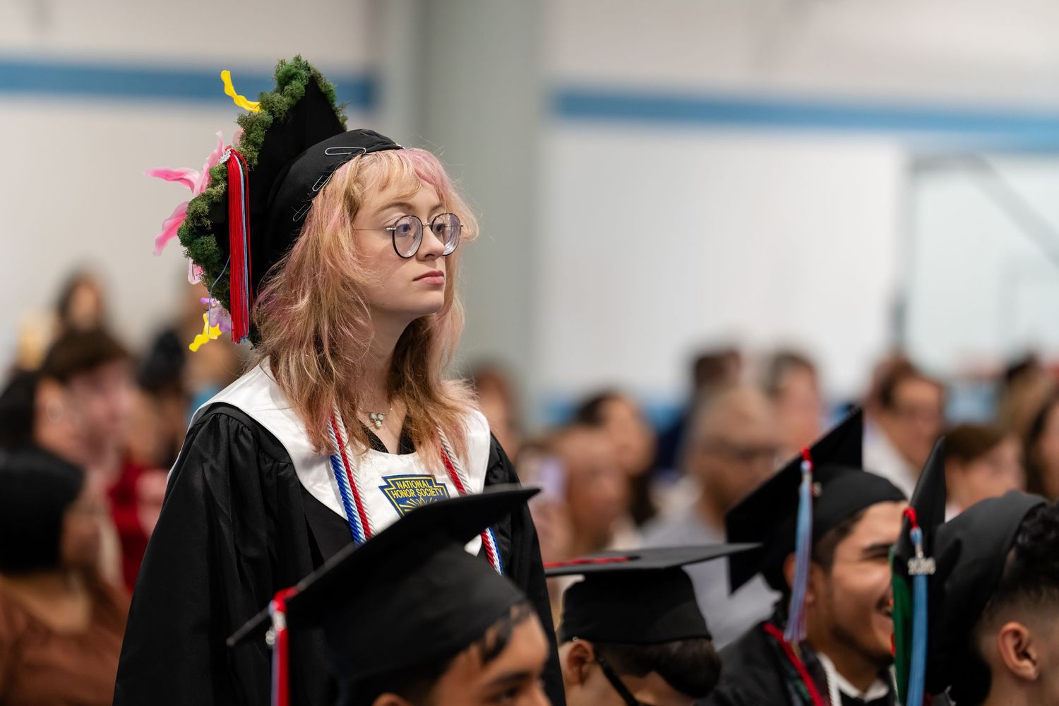 Graduate in decorated cap and gown stands out at a graduation ceremony among seated classmates.