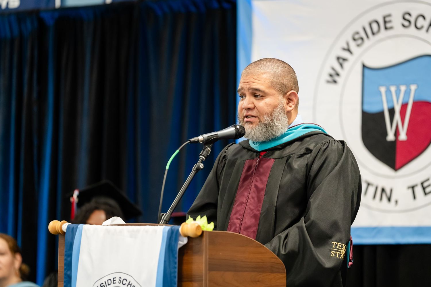 Man in graduation regalia speaks at podium during Wayside School commencement ceremony in Austin, TX.