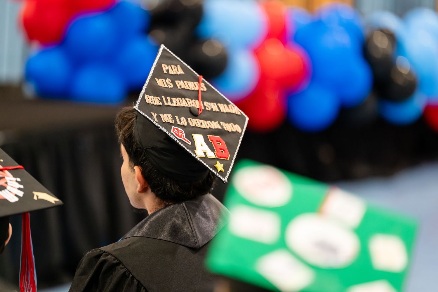 Graduate wearing decorated mortarboard cap with Spanish text at colorful balloon-filled graduation ceremony.