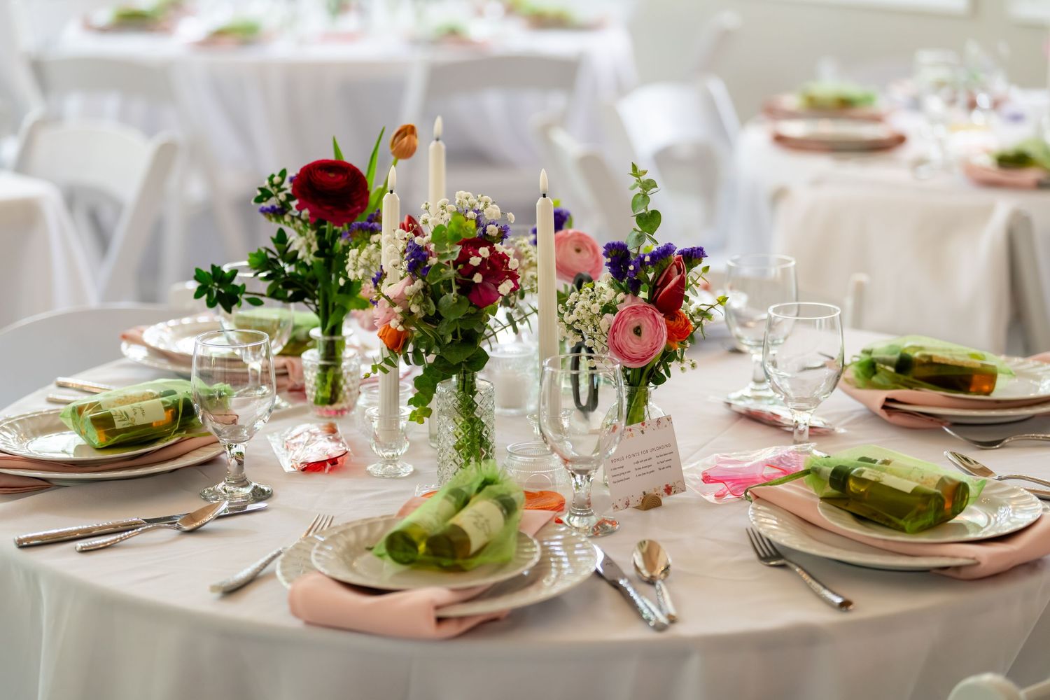 Elegant wedding reception table with colorful floral centerpiece, white linens, crystal glasses, and green napkins on pink plates.