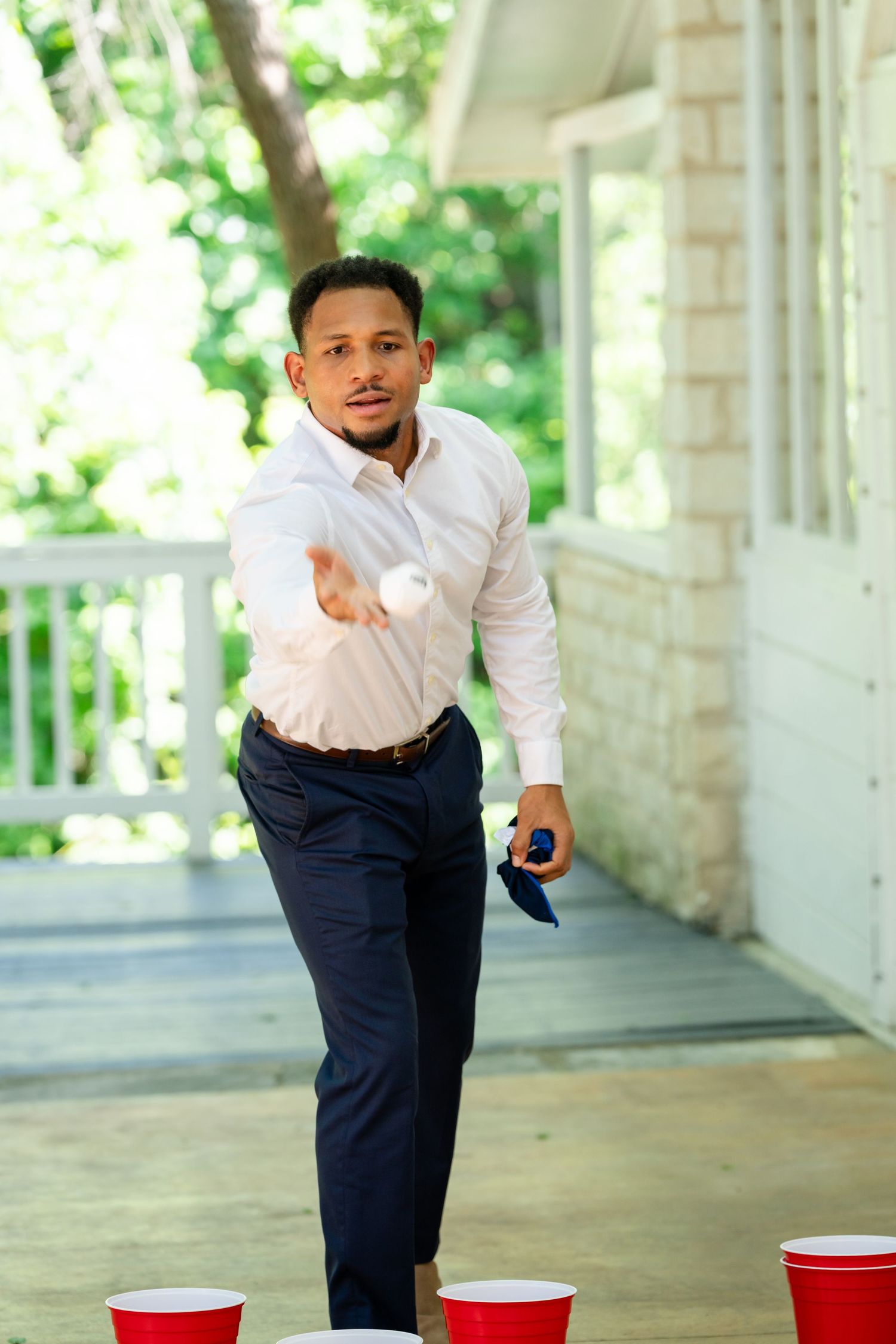 Man in white shirt playing beer pong on a porch, tossing a ping pong ball toward red Solo cups.
