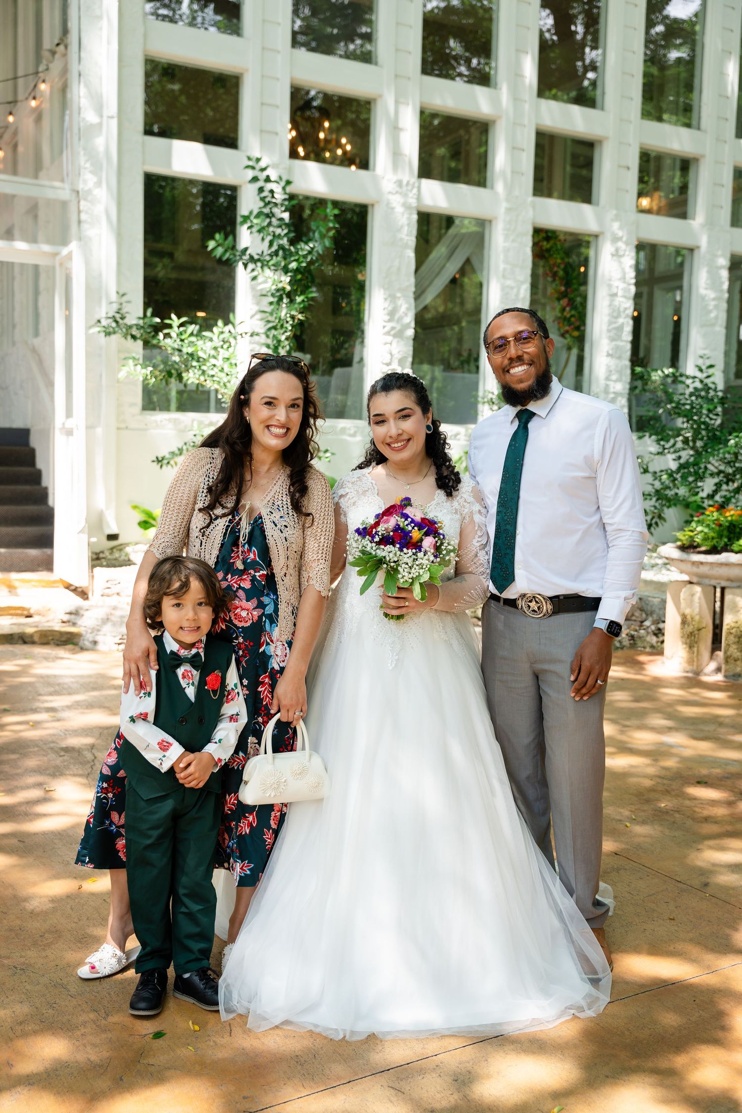 Bride in white gown holds bouquet posing with family including groom in green tie and child at outdoor wedding venue.