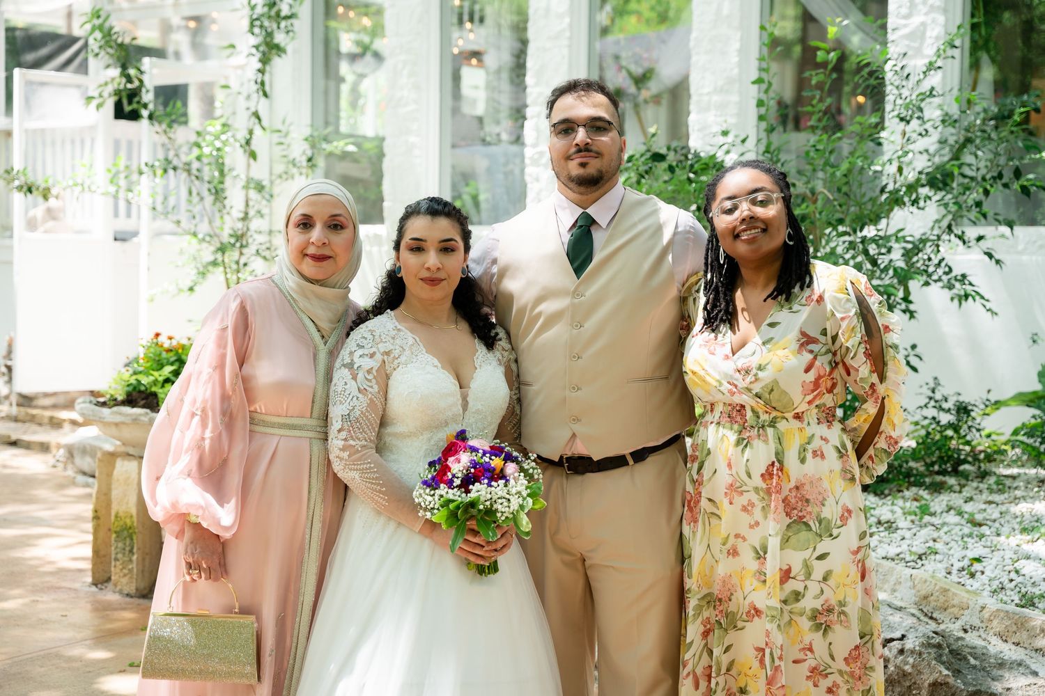 Four guests posing with bride in white wedding gown, wearing elegant formal and floral outfits indoors.