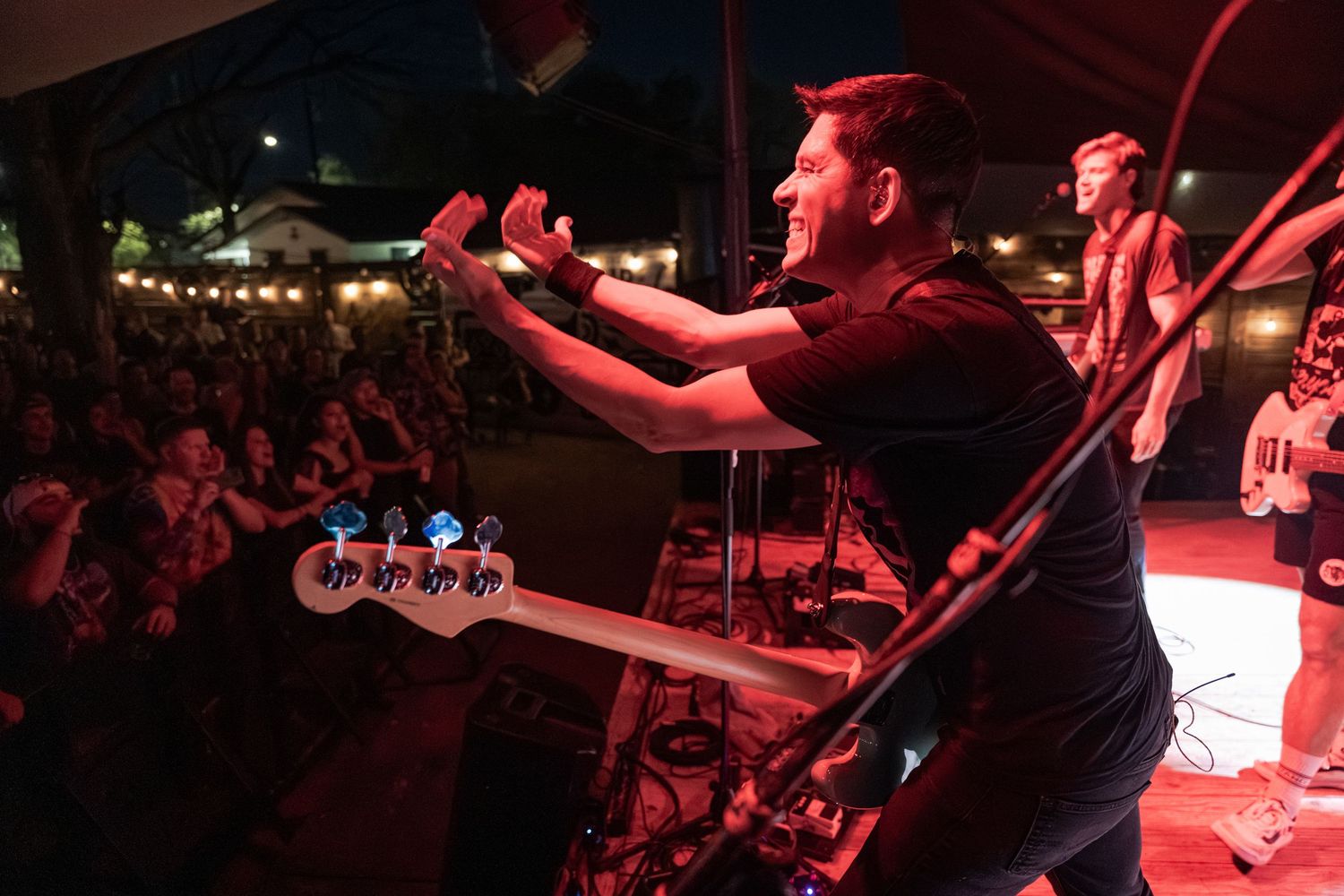 Band performing on stage at night outdoor concert with crowd of fans cheering under string lights.